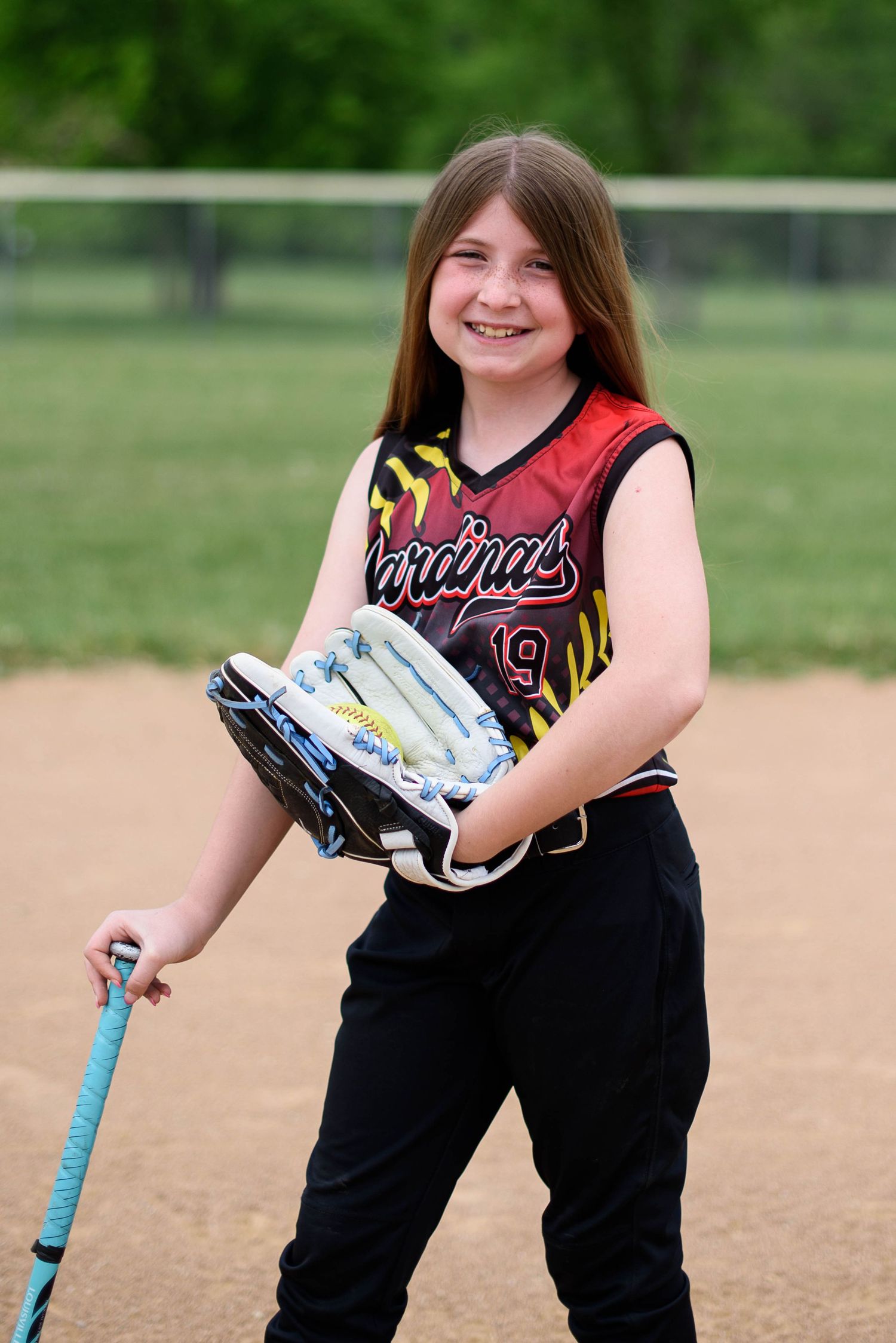 Young girl in Cardinals softball uniform smiling while holding a bat and glove on a baseball field.