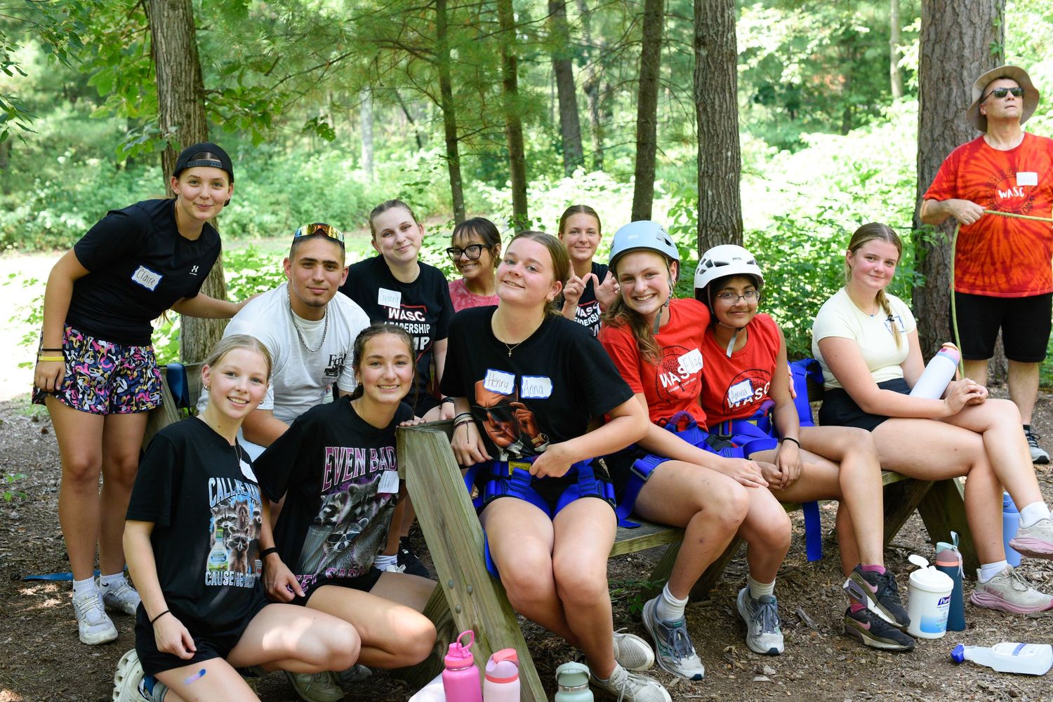 Group of teens and adults smiling together outdoors at a summer camp gathering in a wooded area.