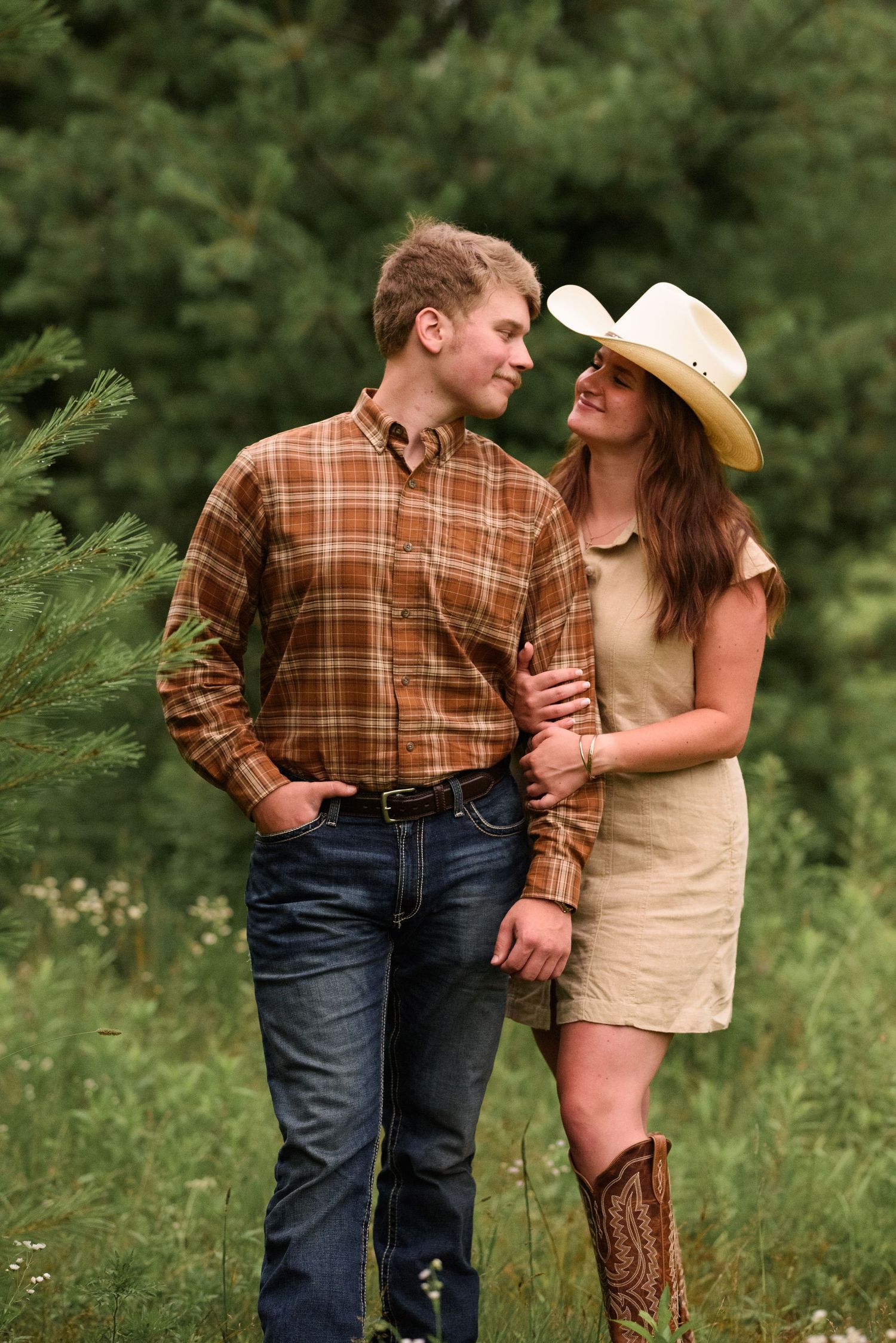 Couple gazing at each other near pine tree, woman in cowboy hat and boots, man in plaid shirt and jeans.
