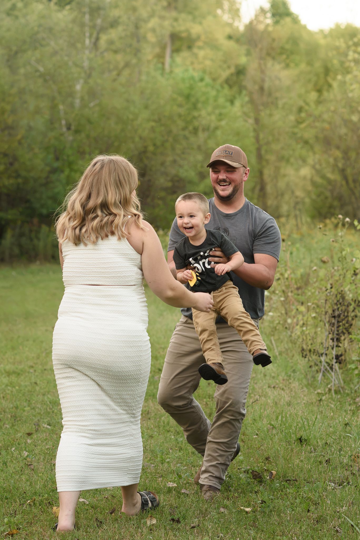 Happy pregnant woman in white dress with man in gray shirt and cap holding laughing toddler outdoors.