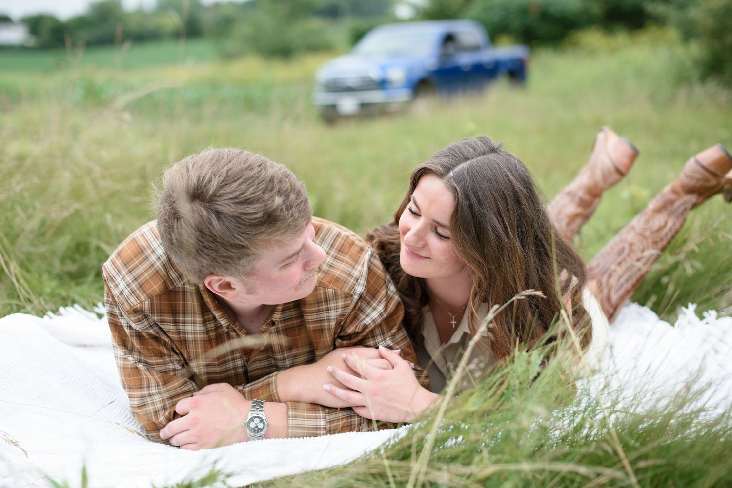 Young couple lying on a white blanket in a grassy field, smiling at each other wearing matching plaid shirts.