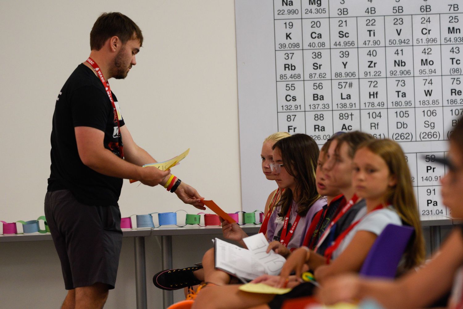 A teacher hands out materials to students in a science classroom with a periodic table on the wall.