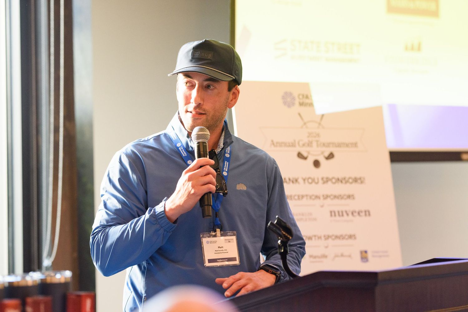 Man in cap speaking at podium with microphone at an annual golf tournament sponsor reception event.