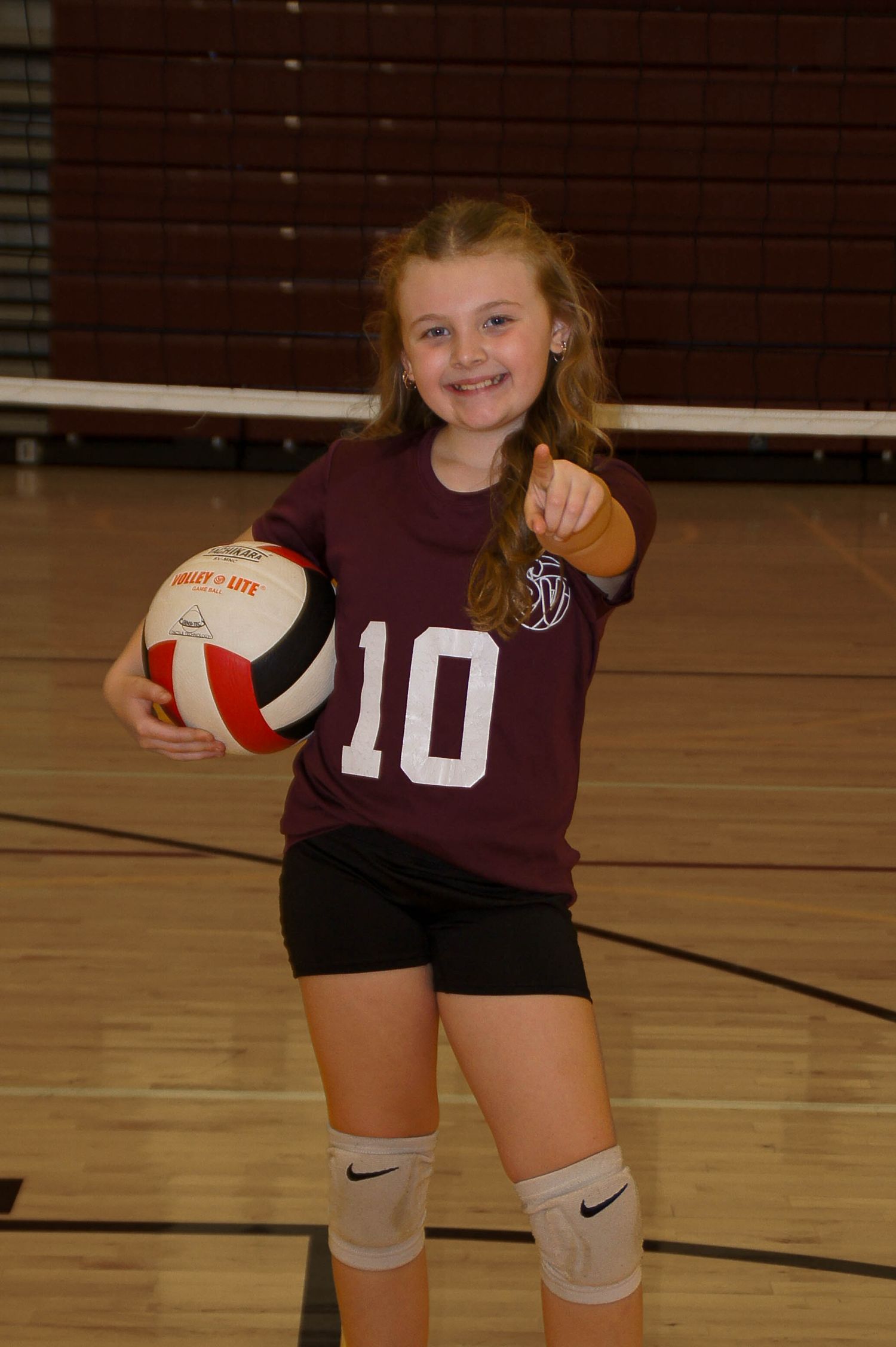 Young volleyball player #10 in a maroon jersey smiles and points while holding a volleyball in a gym.