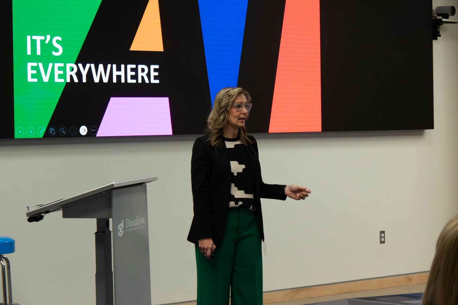 A woman in green pants and black blazer presents at a podium with a colorful 'It's Everywhere' slide behind her.