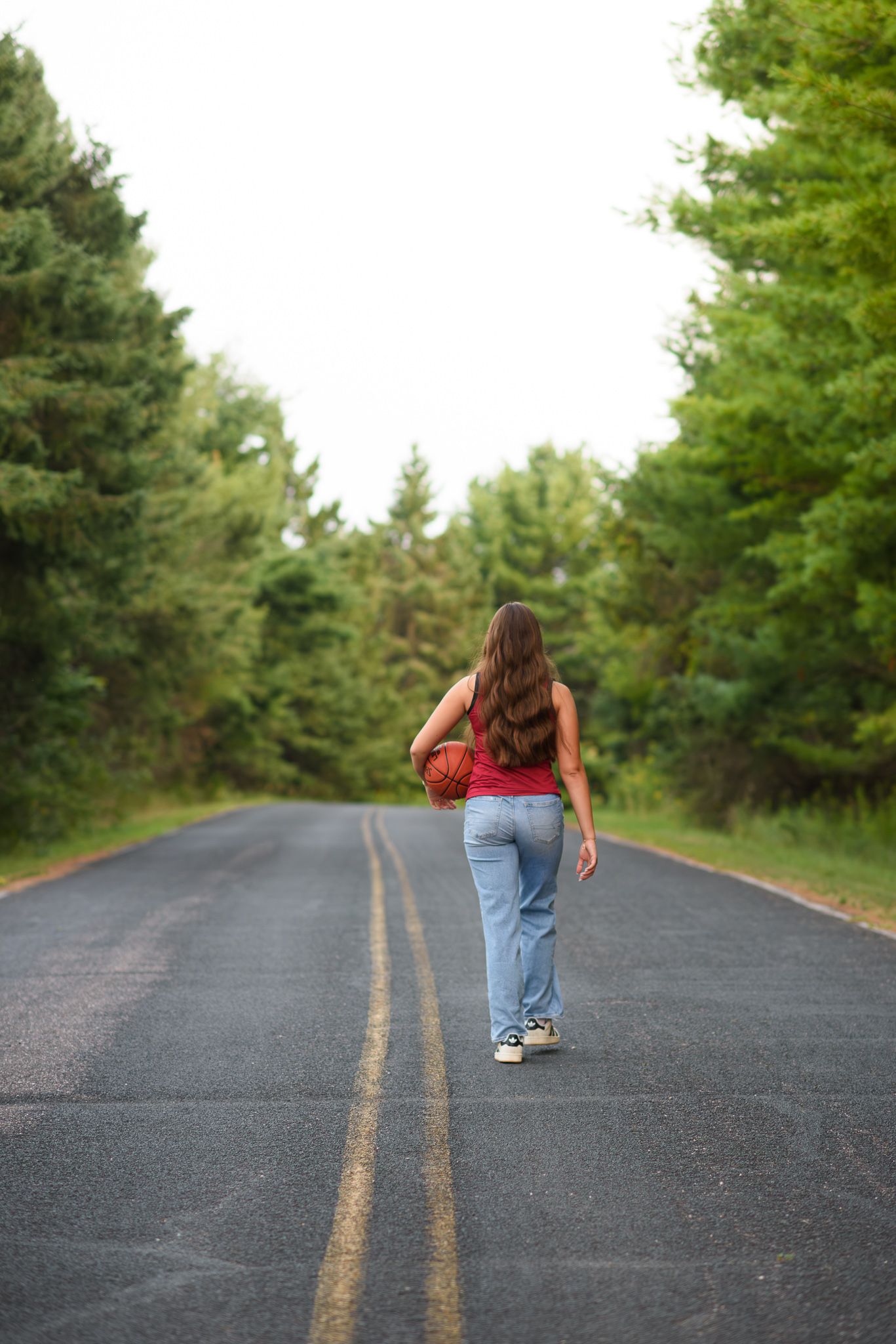 Teen girl in red Cardinals jersey dribbling a basketball while walking down a tree-lined road.