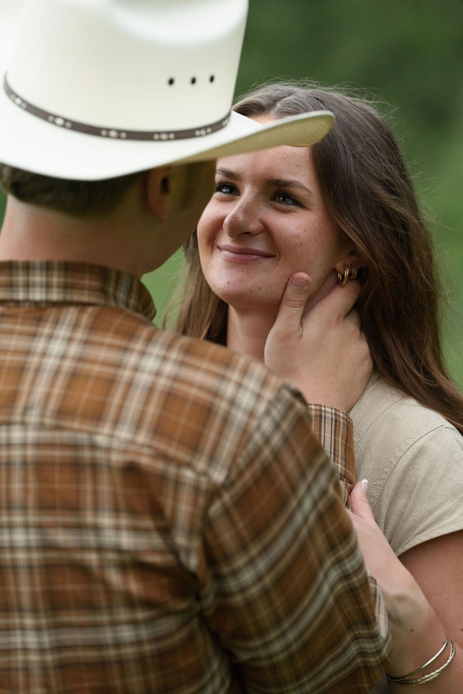 A young woman smiles warmly as a man in a cowboy hat gently touches her face outdoors.