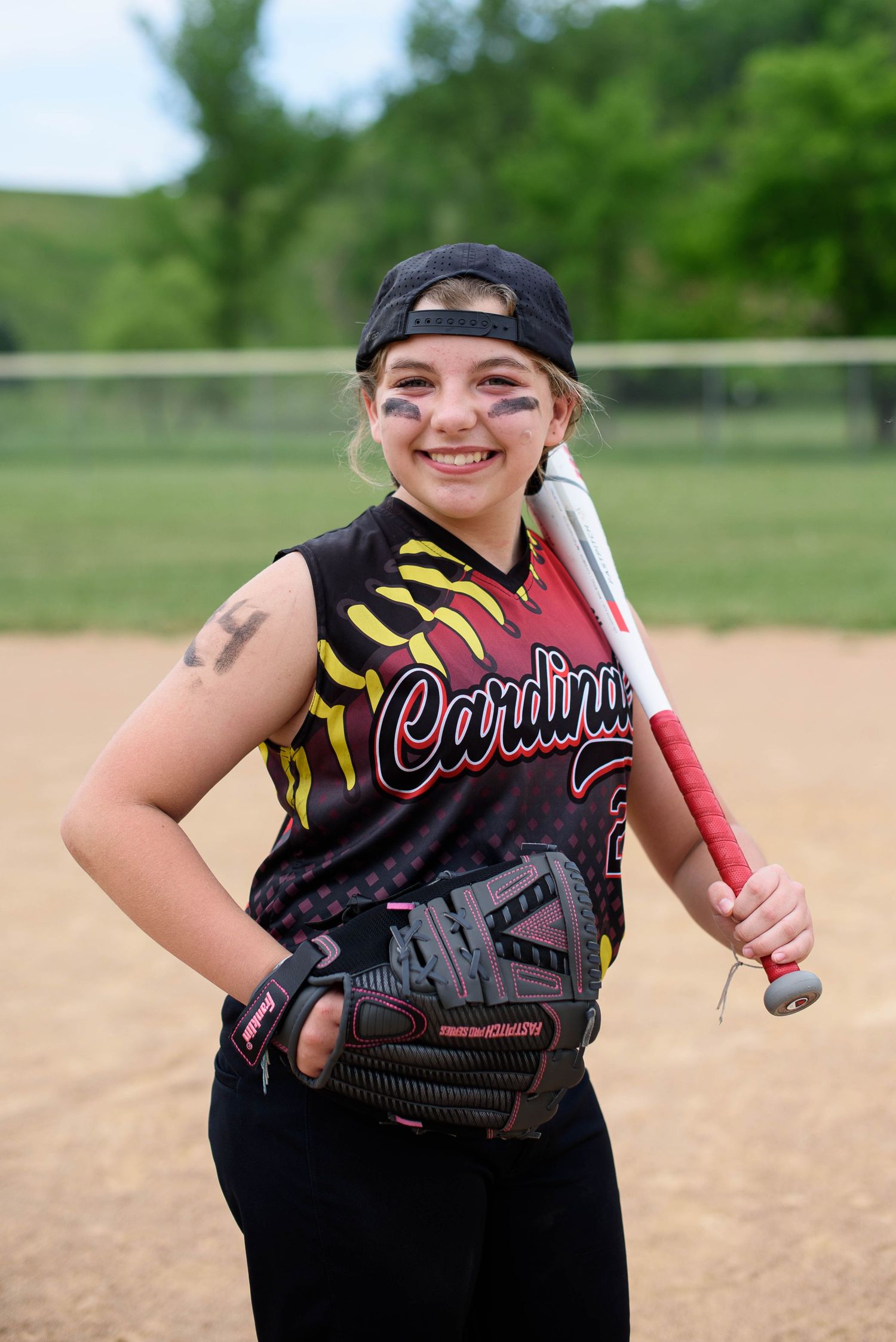 Smiling softball player in Cardinals jersey holds bat and glove with eye black on her cheeks.