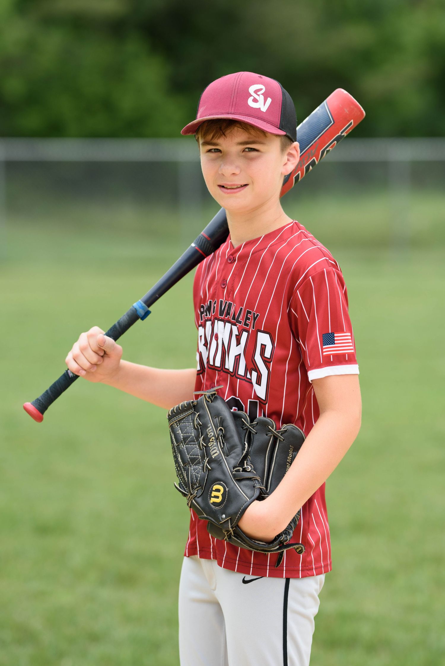 Young SV Cardinals baseball player smiles holding bat on shoulder and glove in red pinstripe uniform outdoors.