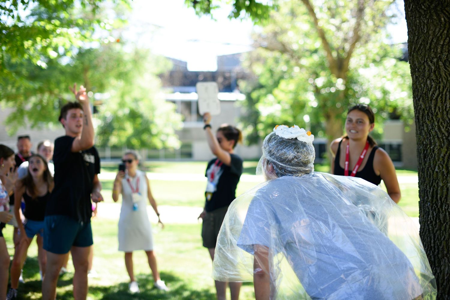 Students play outdoor water activity game on sunny campus lawn, one wearing plastic rain poncho near tree.