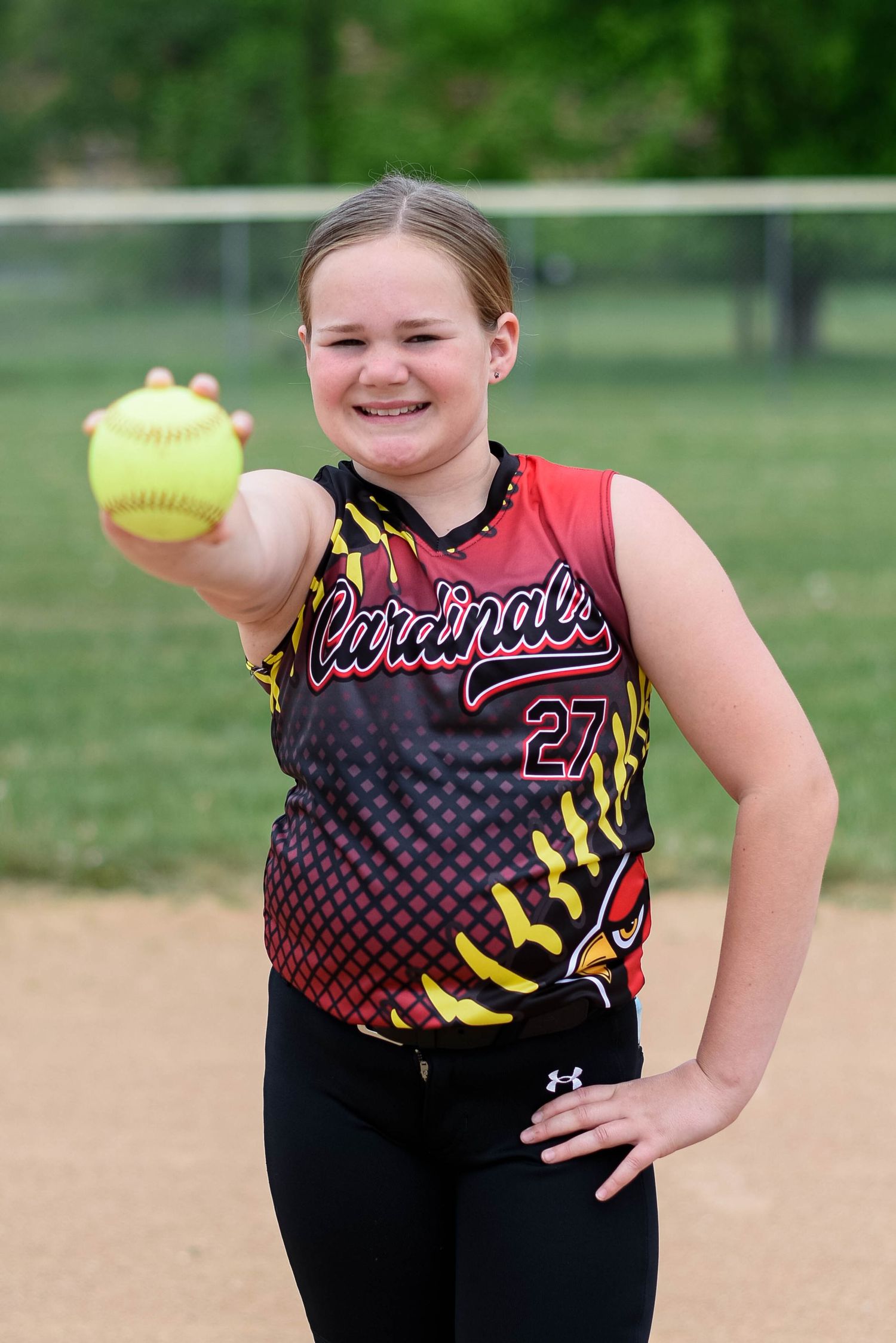 Young Cardinals softball player #27 smiling and holding a yellow softball toward the camera on a dirt field.