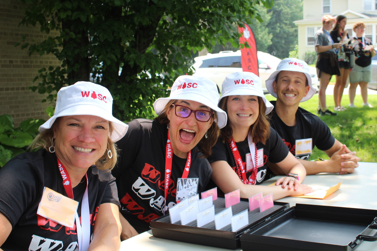 Four smiling women wearing W&SC hats sit at an outdoor registration table during a sunny campus event.