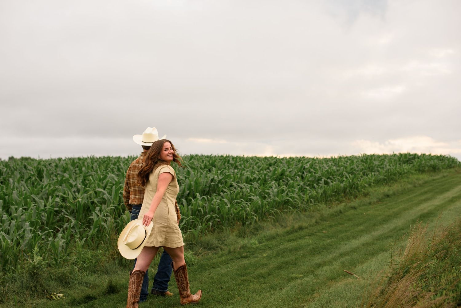 Two women in cowboy boots and hats walk along a lush green cornfield path under an overcast sky.