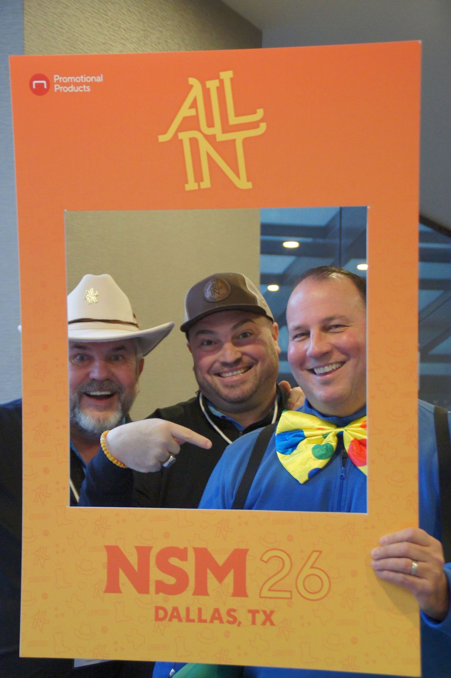 Three men in costumes smile through an All In NSM 26 Dallas TX promotional photo frame.