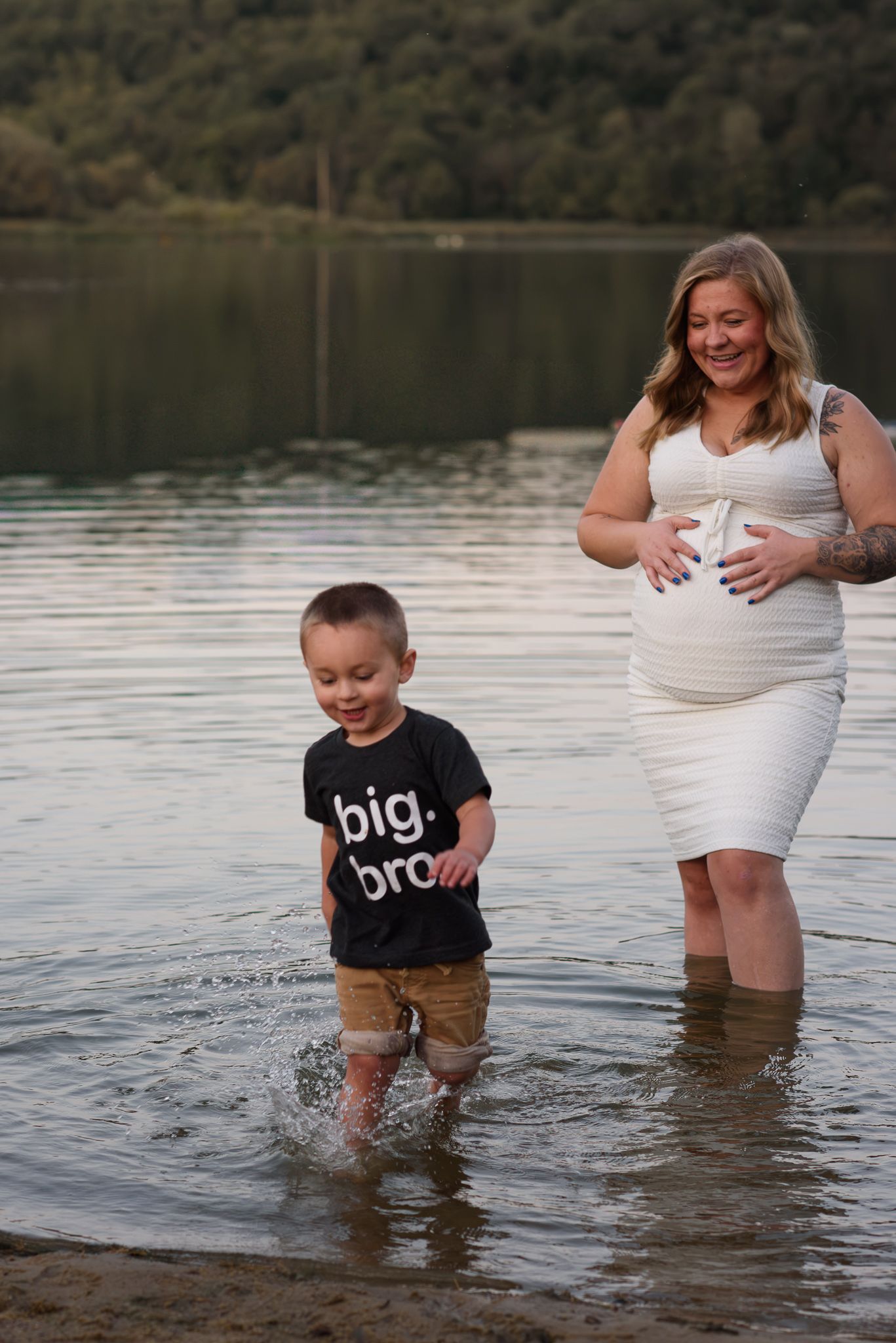 Pregnant woman in white dress with toddler in big bro shirt splashing in lake.