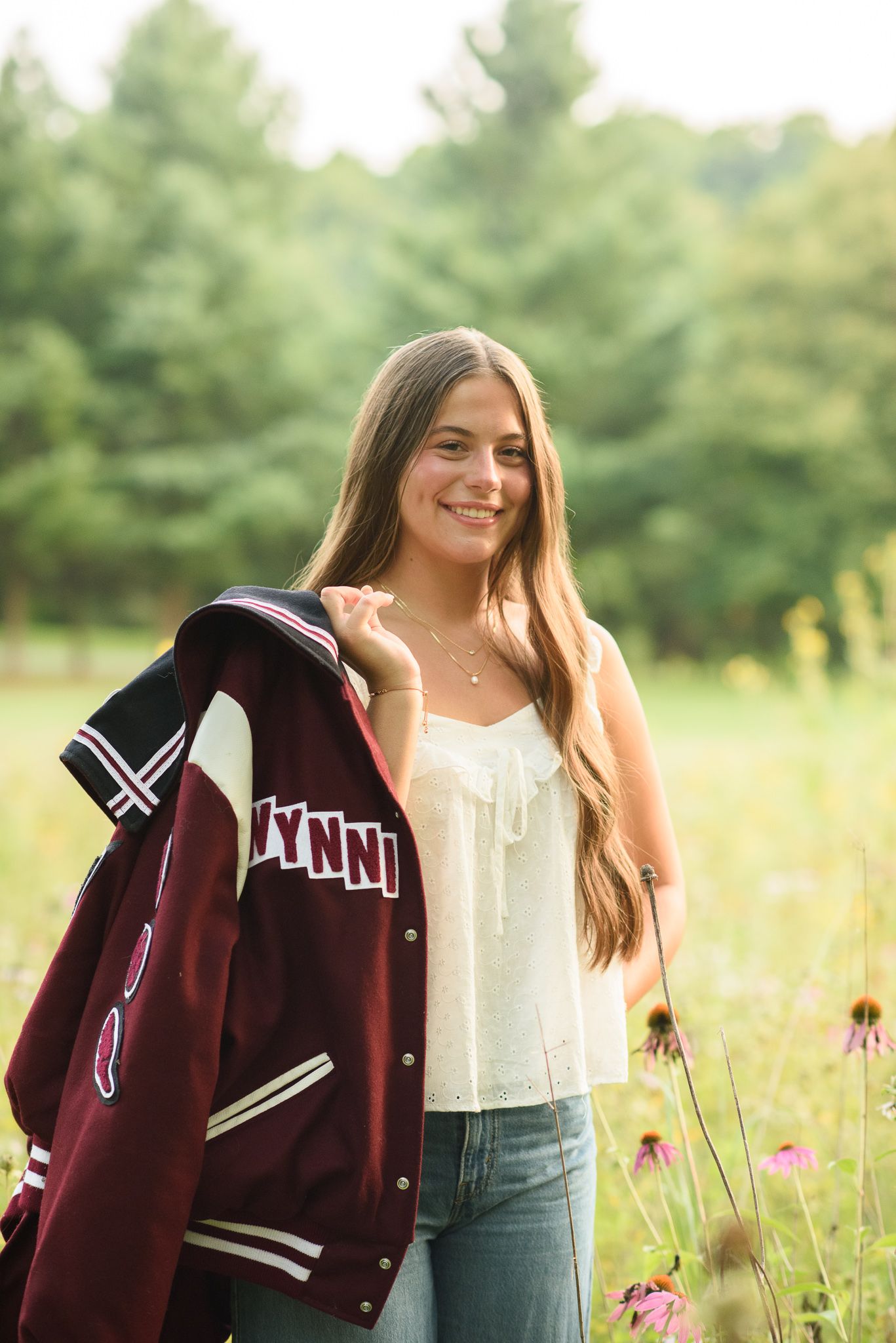 High school senior girl holding maroon letterman jacket in flower field wearing white top and jeans.