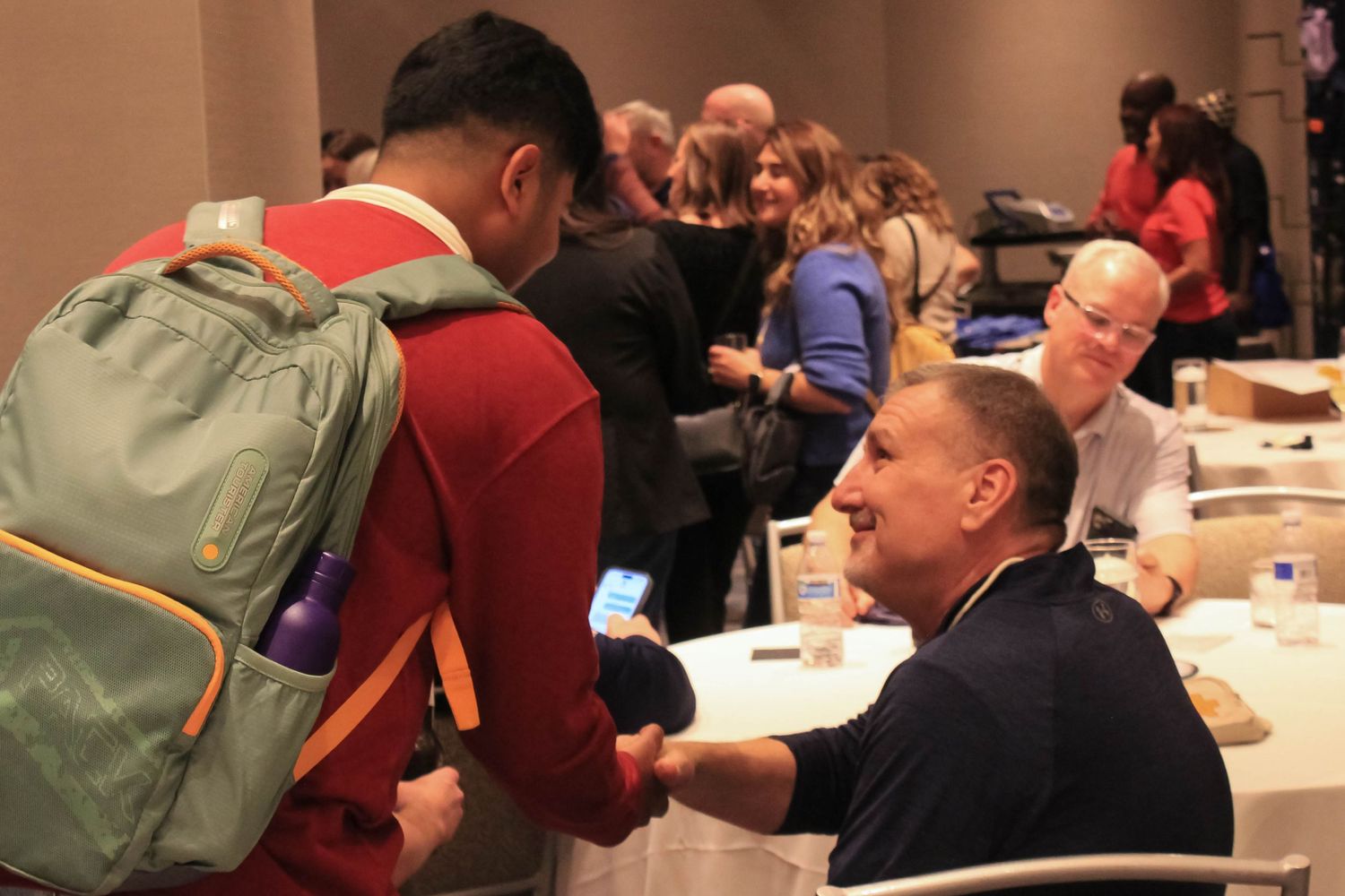 Man with backpack showing phone to seated man at a conference event.