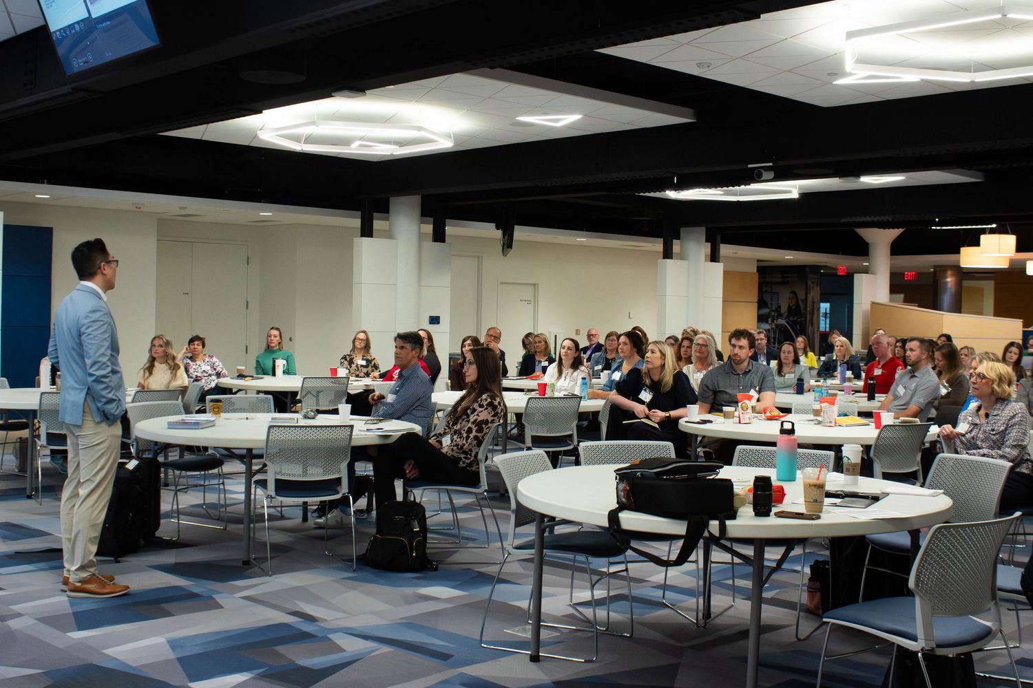 A presenter speaks to a large group of attendees seated at round tables during a professional conference event.