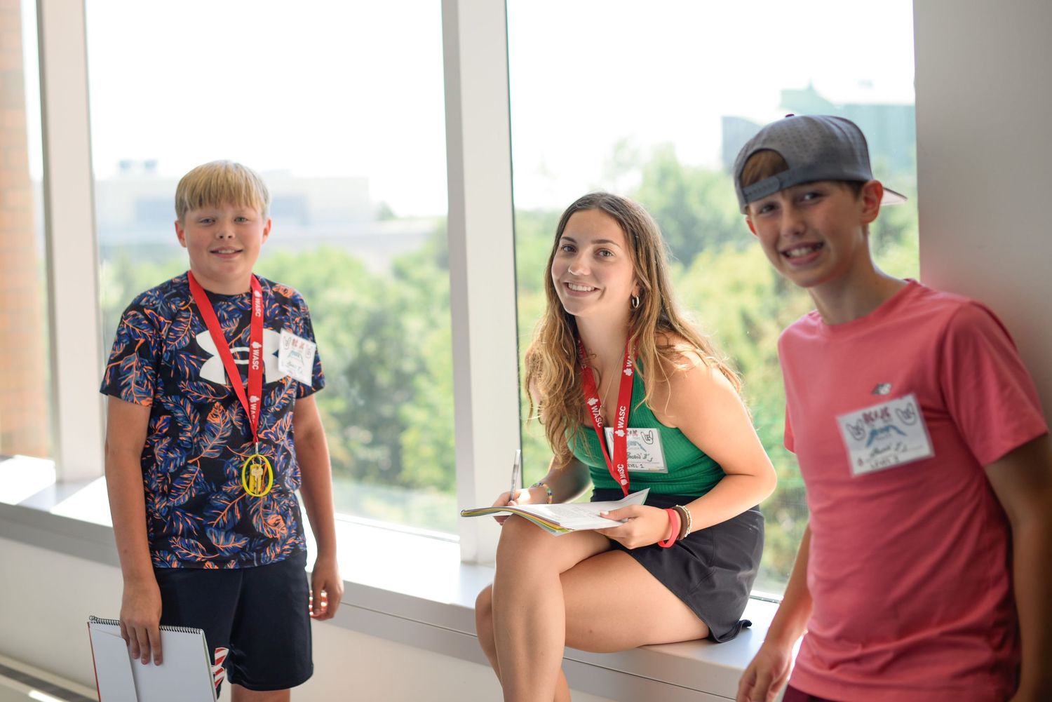 Three smiling kids wearing lanyards and name tags pose together near a bright window at a summer camp event.