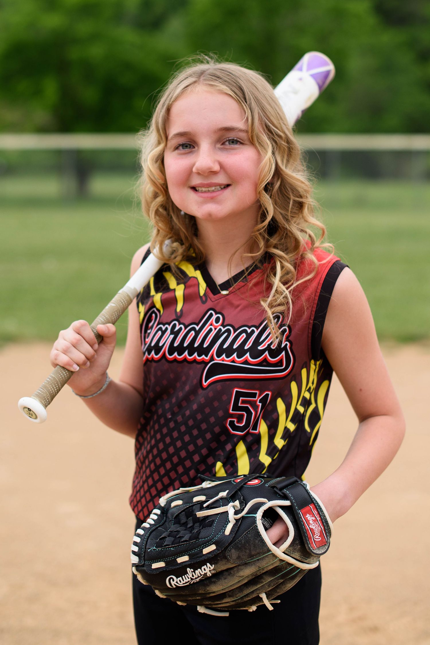 Young softball player in Cardinals #51 jersey poses with bat and Rawlings glove on a baseball diamond.