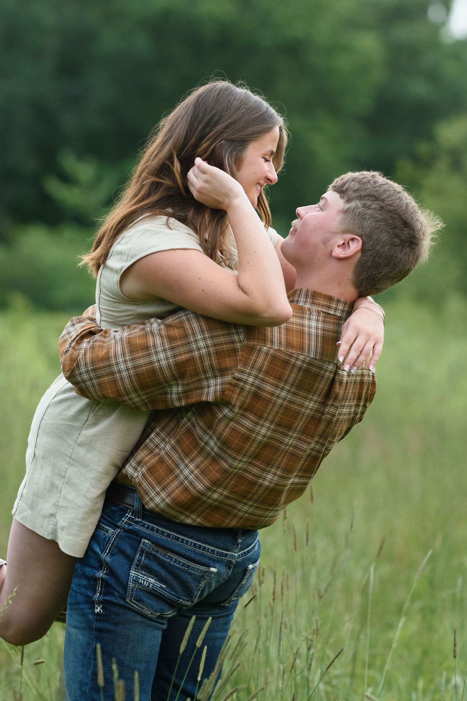 Man in brown plaid shirt lifting woman in green field, both laughing joyfully in romantic outdoor moment.
