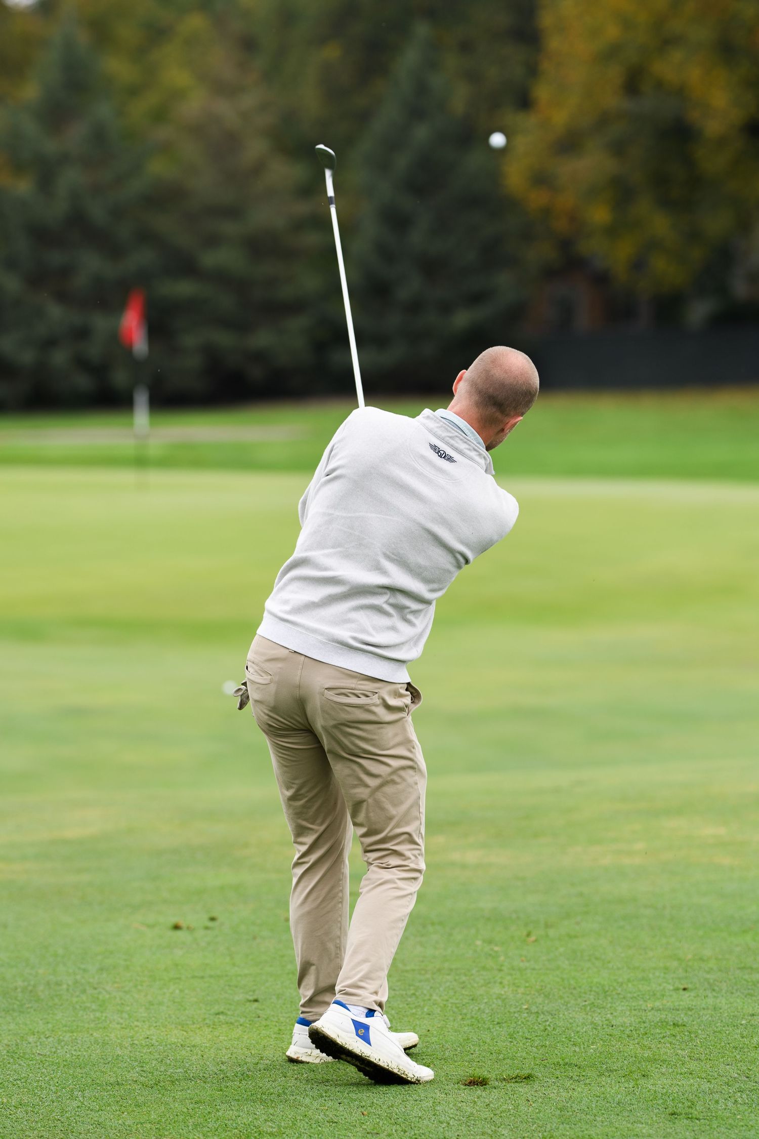 Bald golfer in white shirt and khaki pants completing a golf swing on a lush green course.