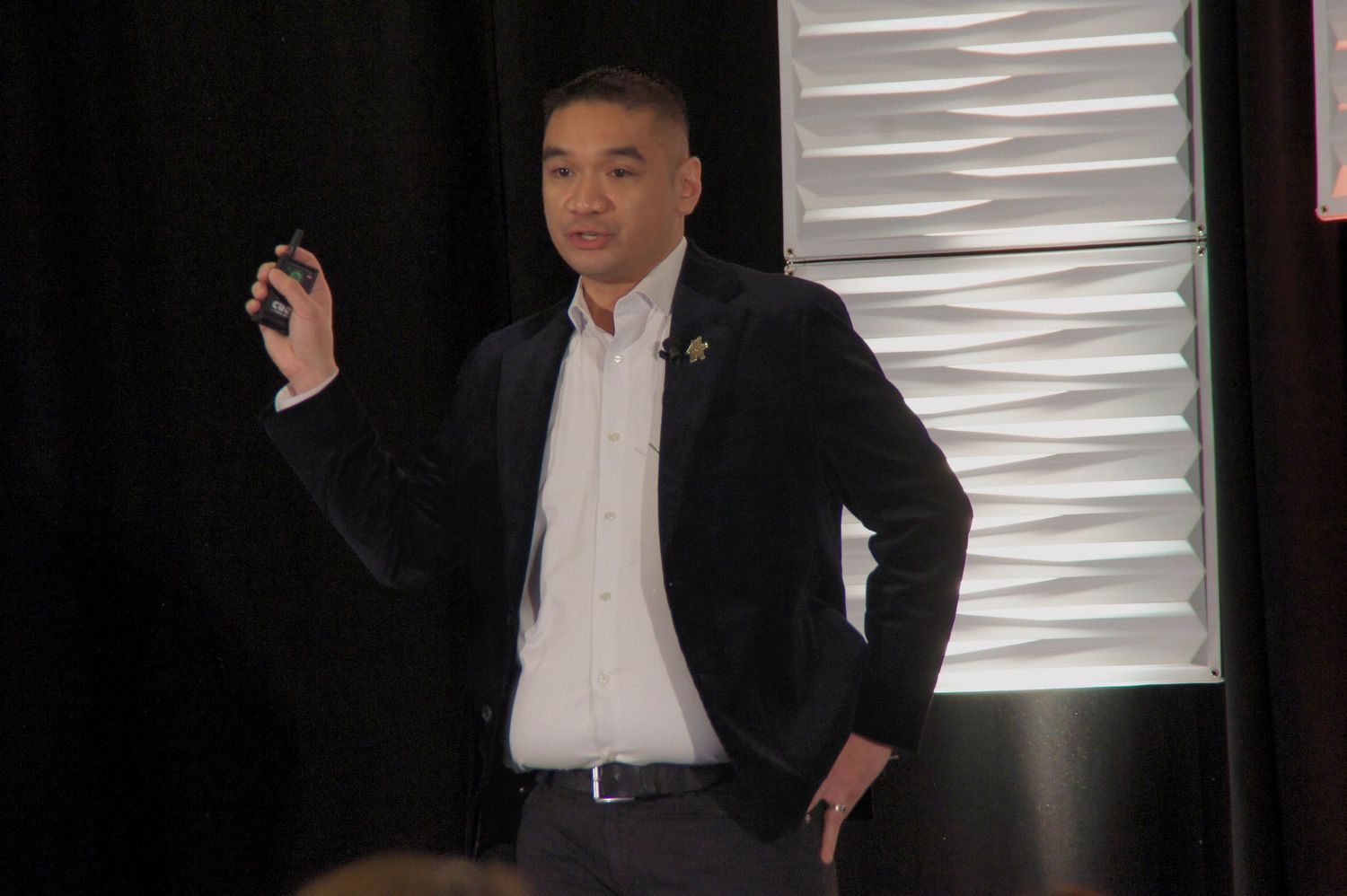 A man in a black blazer presents at a conference, holding a remote clicker while speaking on stage.
