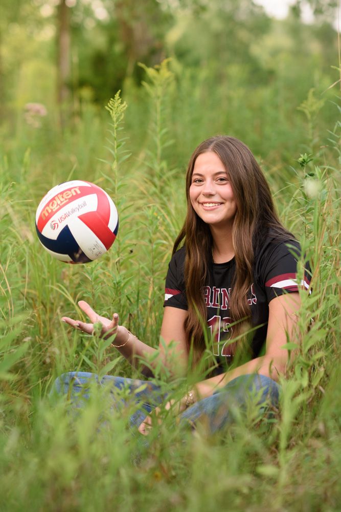 A smiling teenage girl in a volleyball jersey sits in tall green grass, tossing a red, white, and blue volleyball in the air.