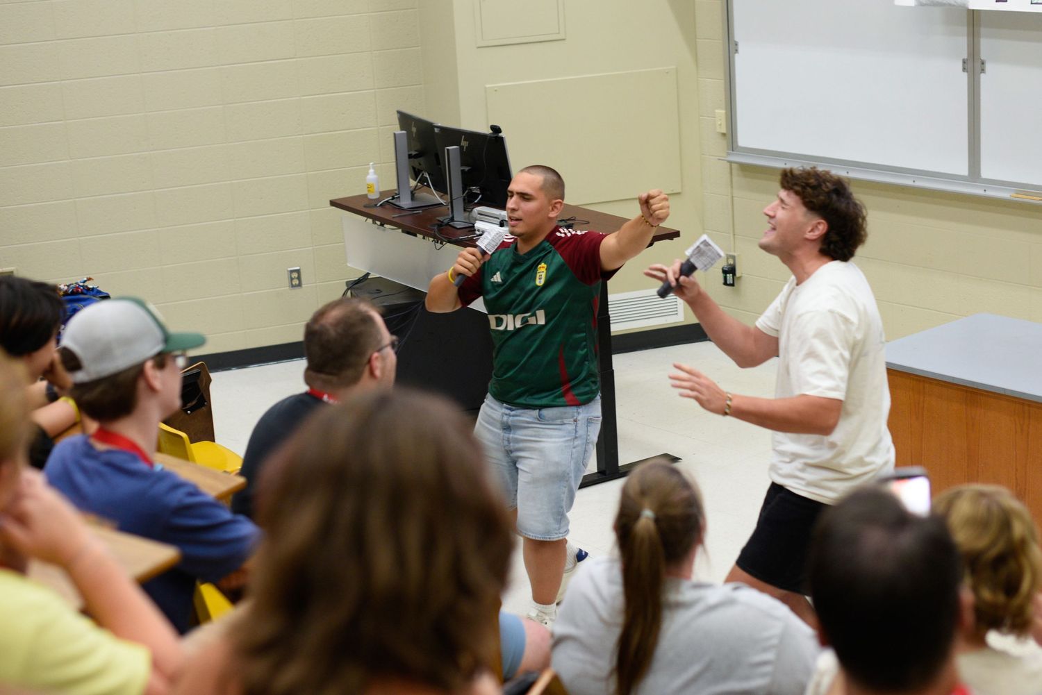 Two men engage animatedly with students in a college lecture hall classroom setting.