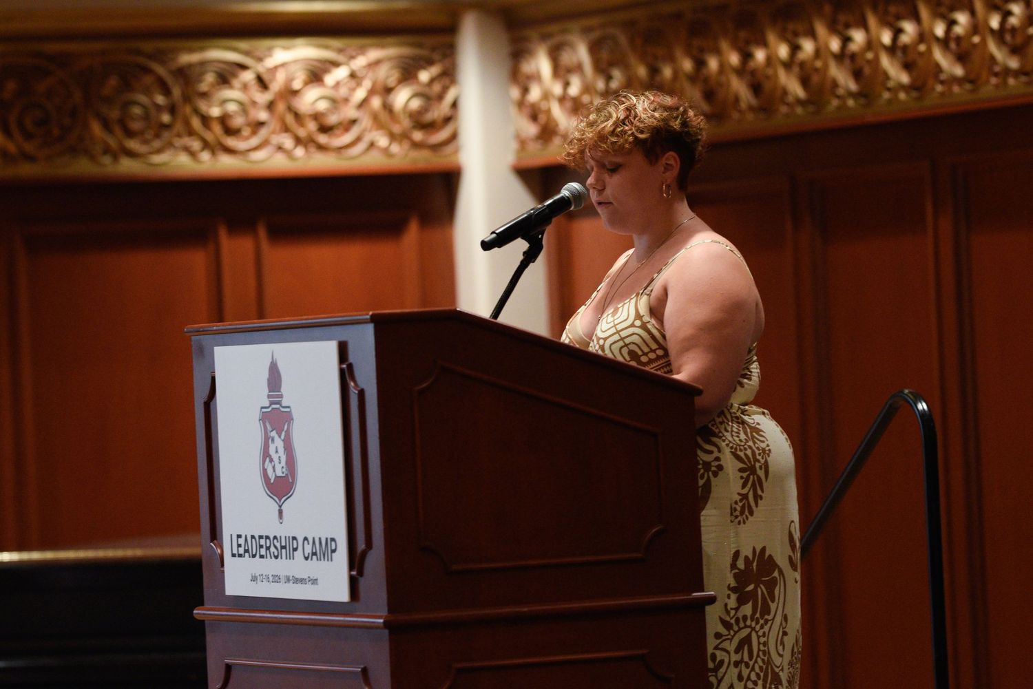 Woman speaking at podium during Leadership Camp event in ornate venue.