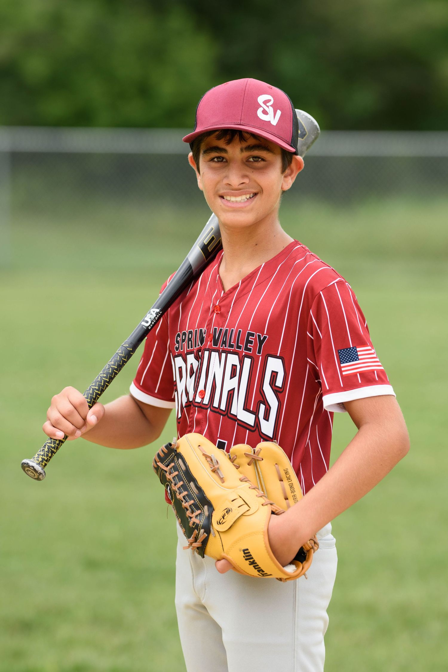 Young baseball player in Spring Valley Cardinals uniform smiling, holding bat over shoulder and wearing fielding glove.