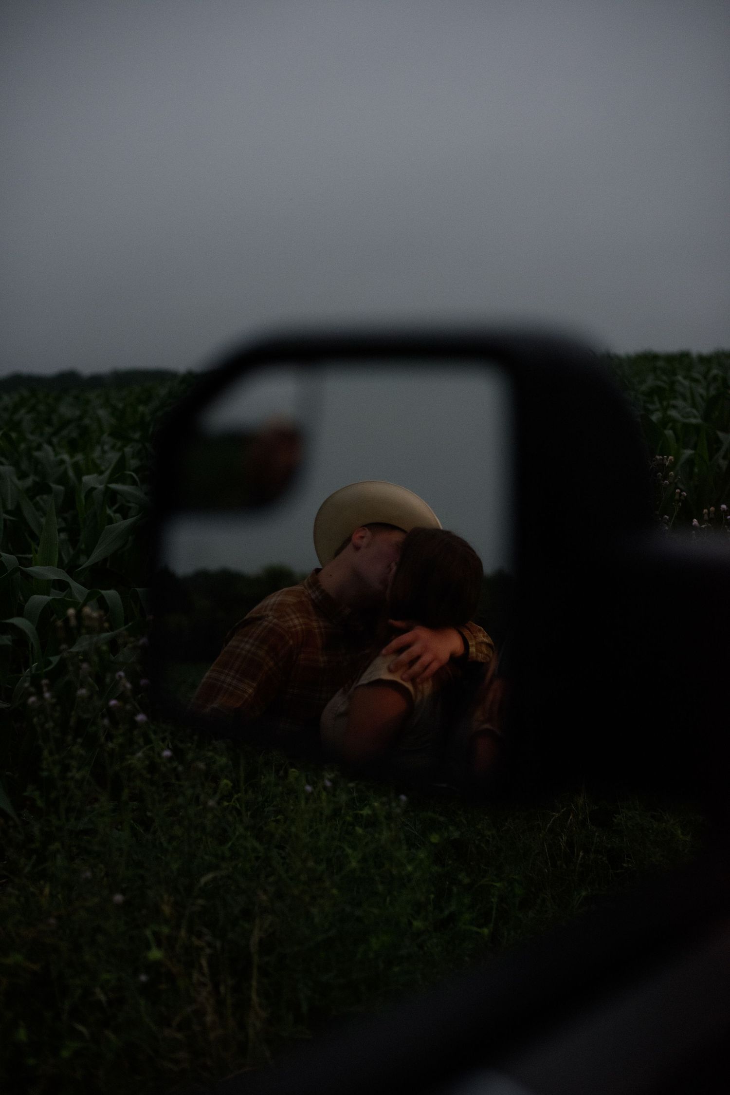 Romantic couple kissing near cornfield reflected in truck side mirror during overcast evening.