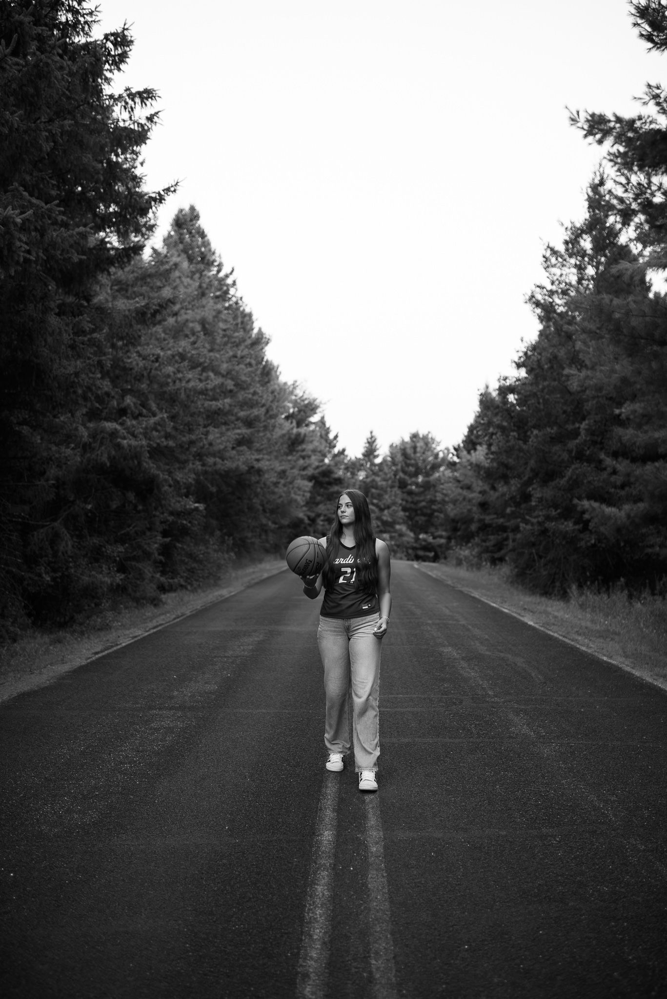 Black and white photo of a young woman holding a basketball standing on a tree-lined road.
