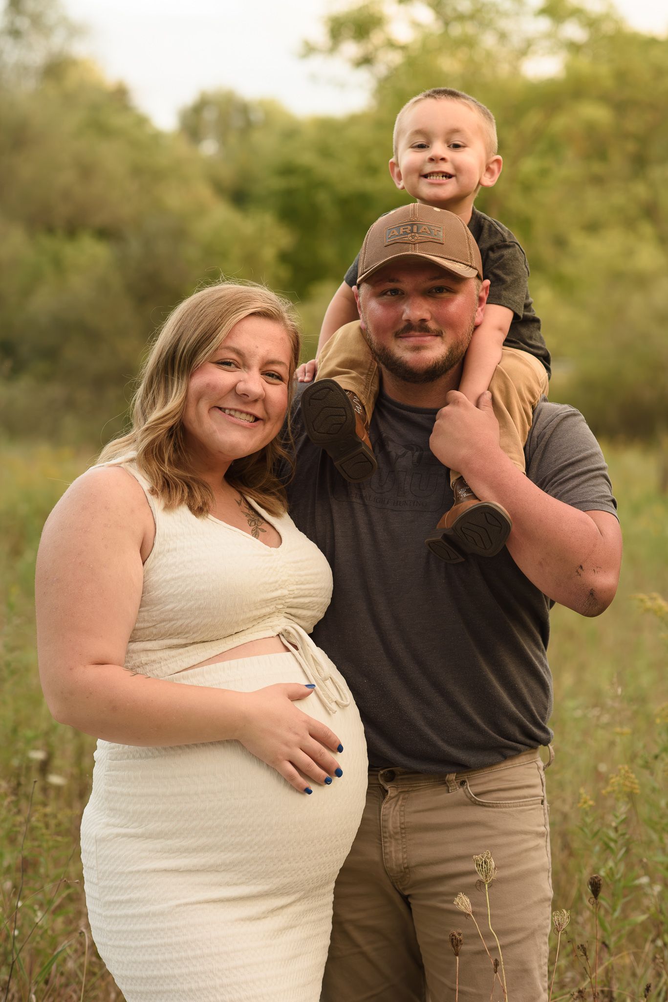 Pregnant woman in white dress poses with husband and young son on his shoulders in an outdoor field setting.