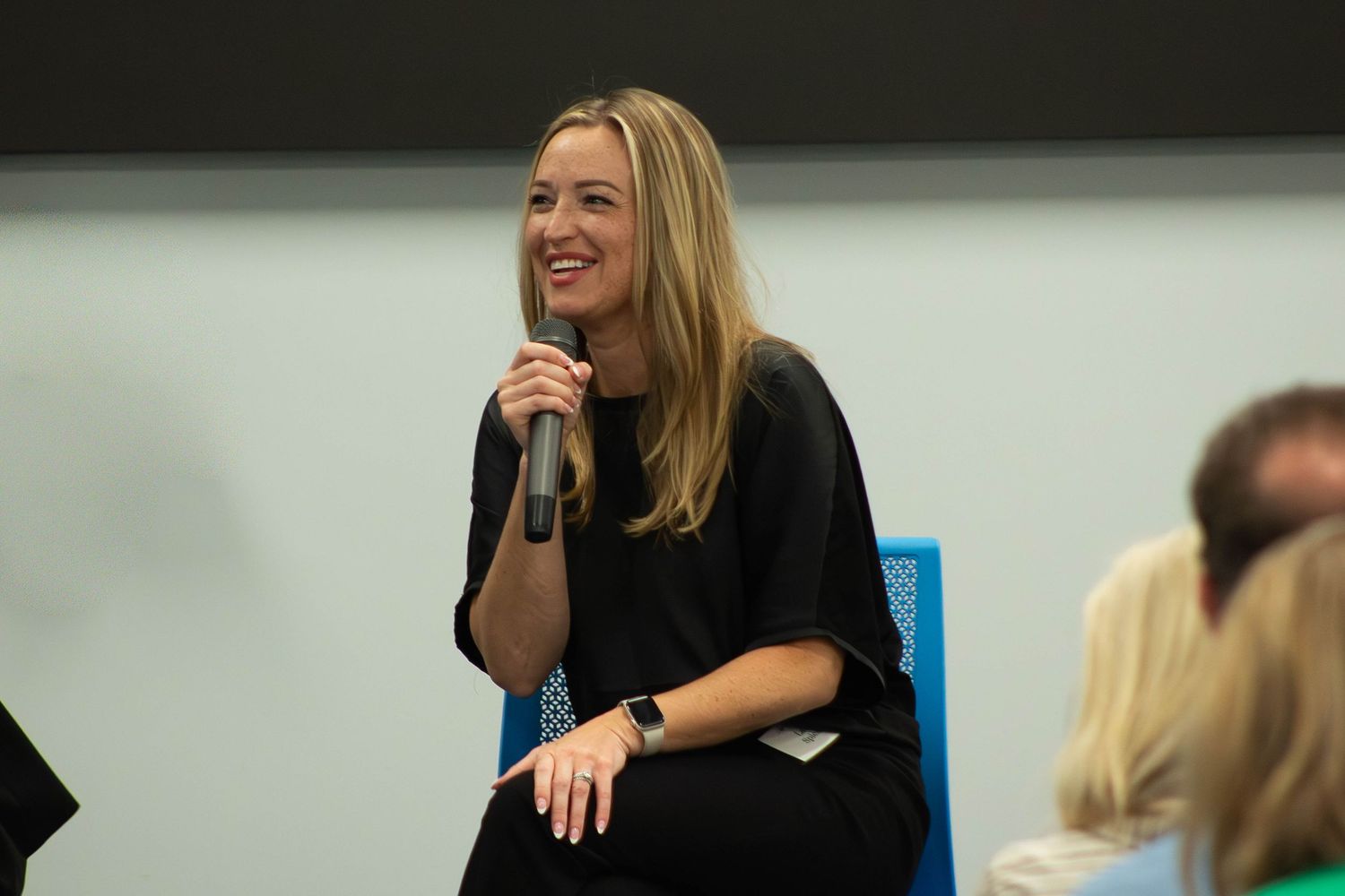 A smiling woman in a black outfit holds a microphone while seated at a professional panel discussion event.