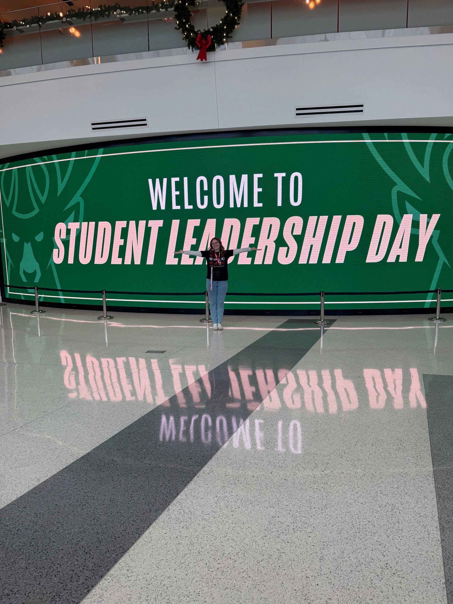 Student stands before Milwaukee Bucks Welcome to Student Leadership Day banner with reflective floor.