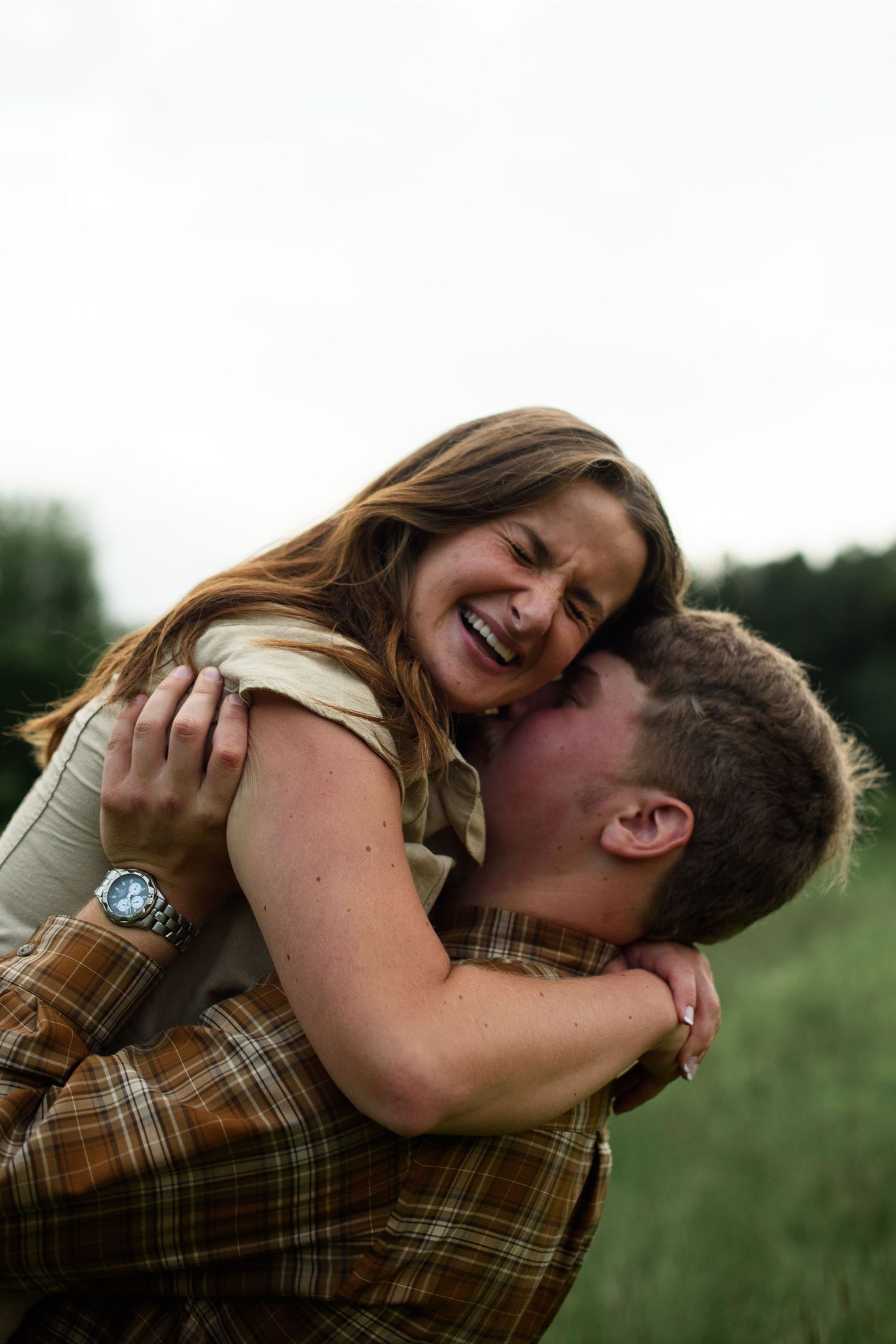 A joyful couple embraces in a green field, woman laughing as man hugs her, both wearing brown plaid outfits.