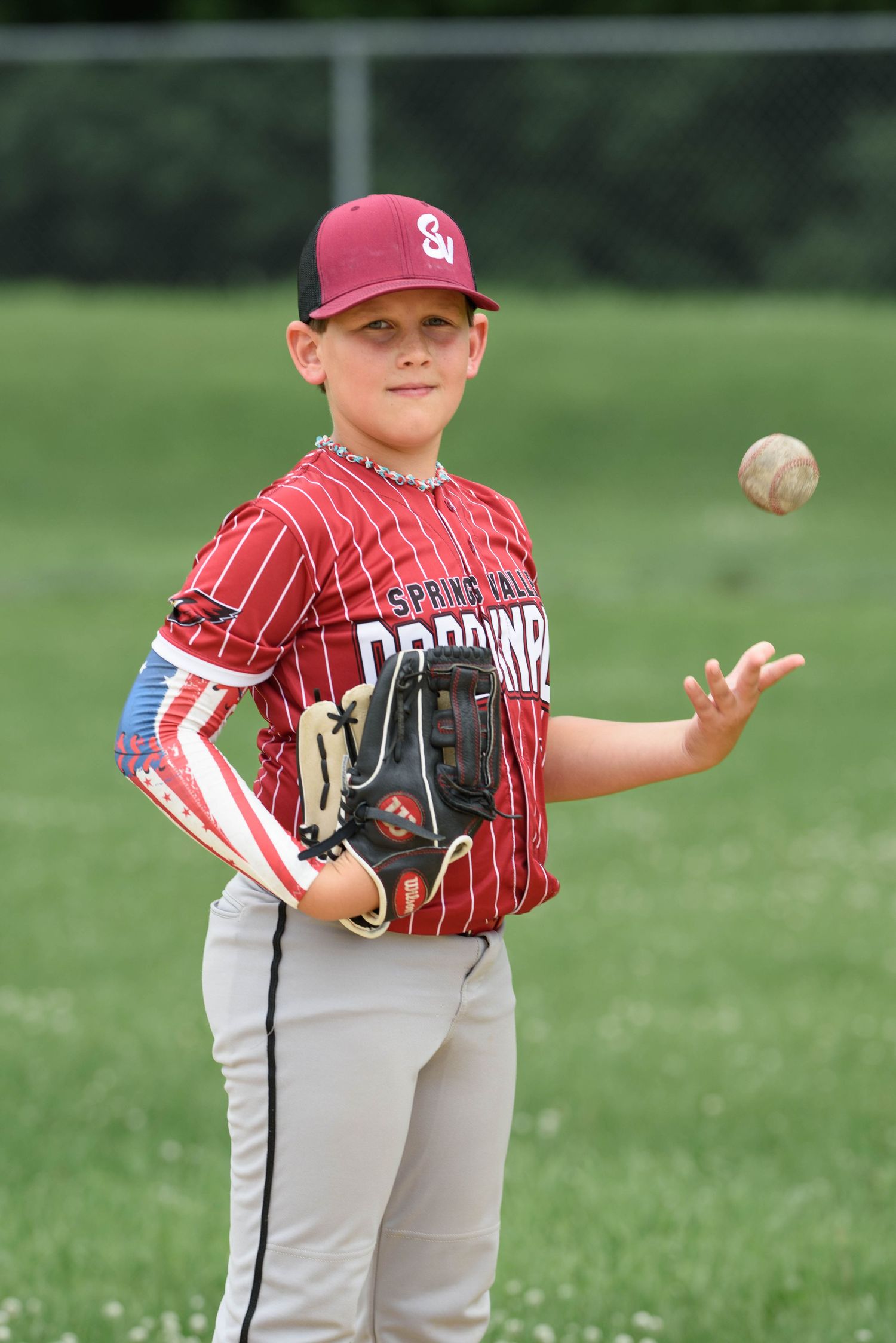 Young baseball player in red Spring Valley Cardinals uniform tossing a ball while wearing a glove on a green field.