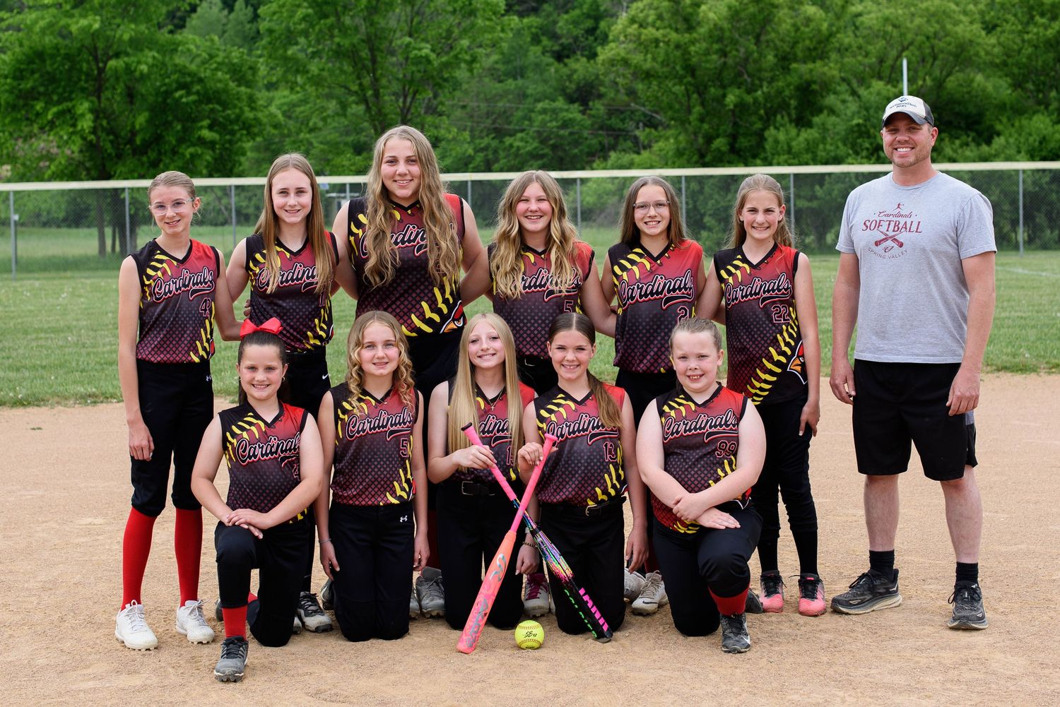 Youth girls softball team poses in black and red uniforms with bats and equipment on a green field with coach.