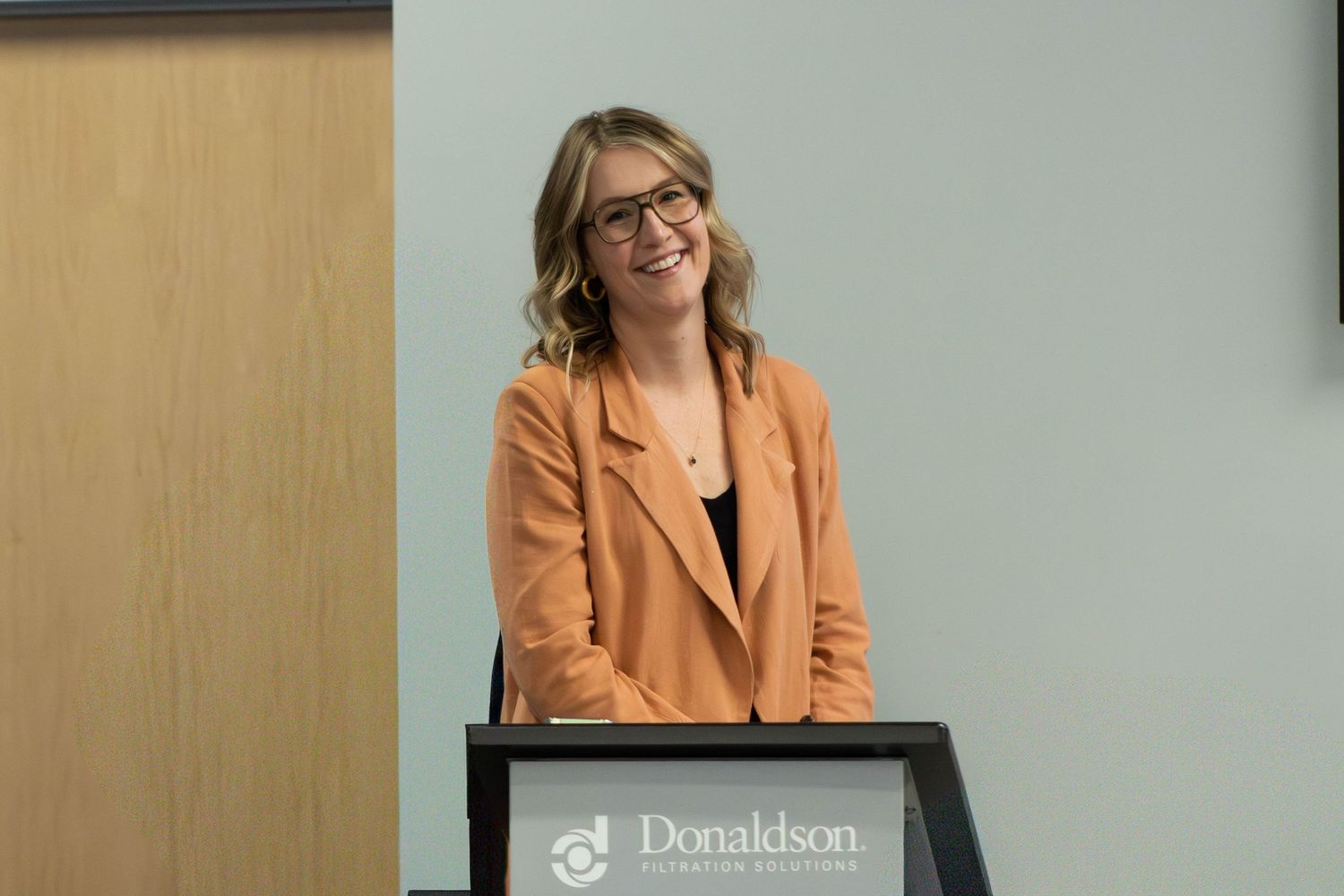 A smiling woman in an orange blazer stands at a Donaldson branded podium during a corporate presentation.