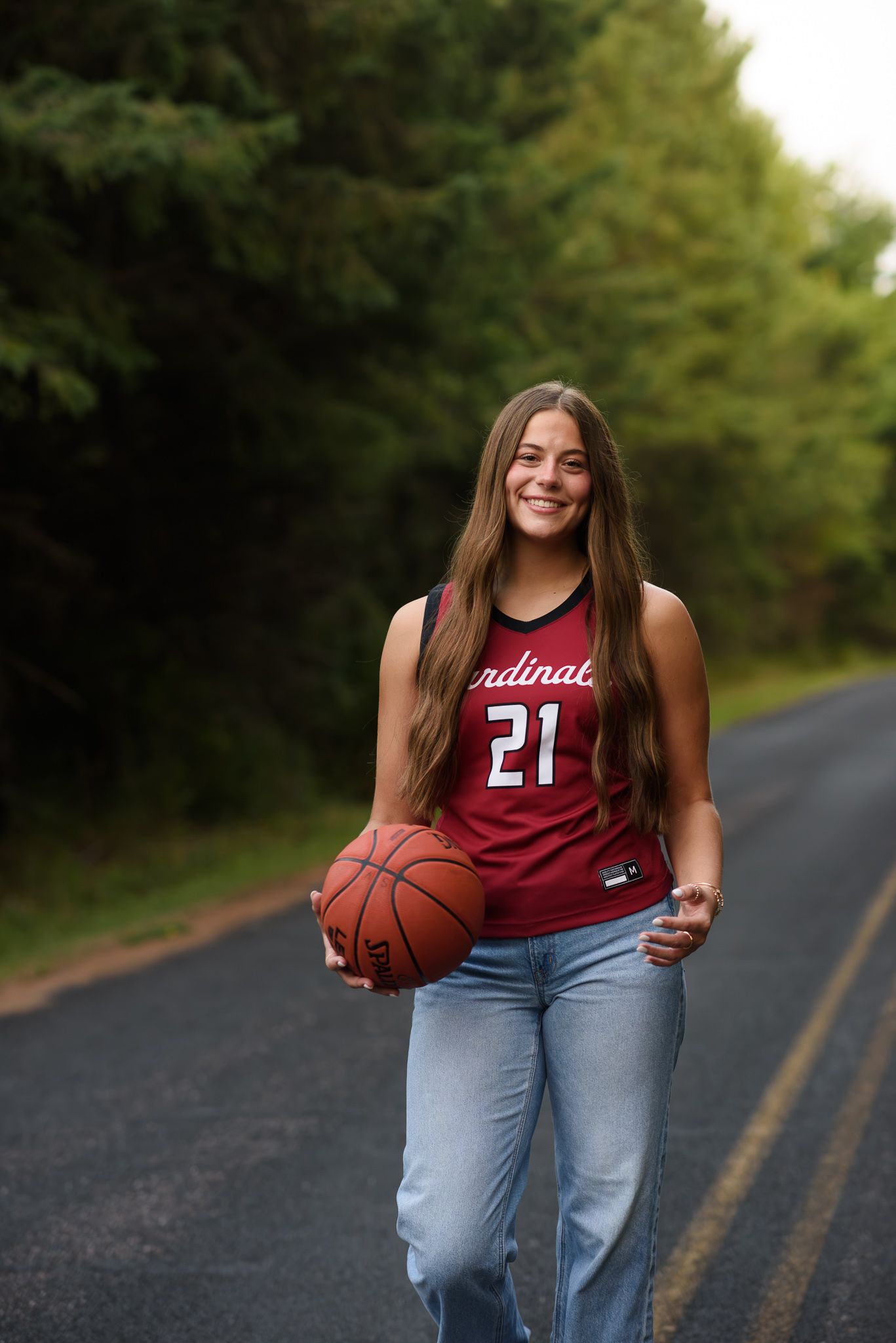 A smiling girl in a Cardinals #21 basketball jersey holds a basketball while walking on a tree-lined road in jeans.