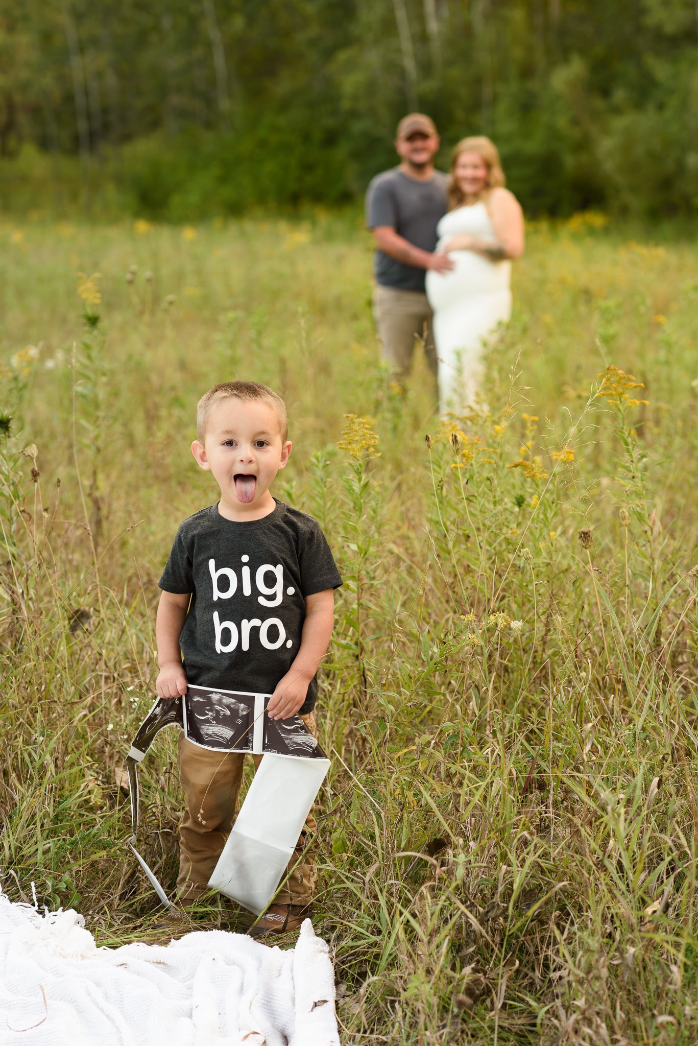 Young boy in 'big bro' shirt holds ultrasound photo as expecting parents embrace in a sunny meadow behind him.