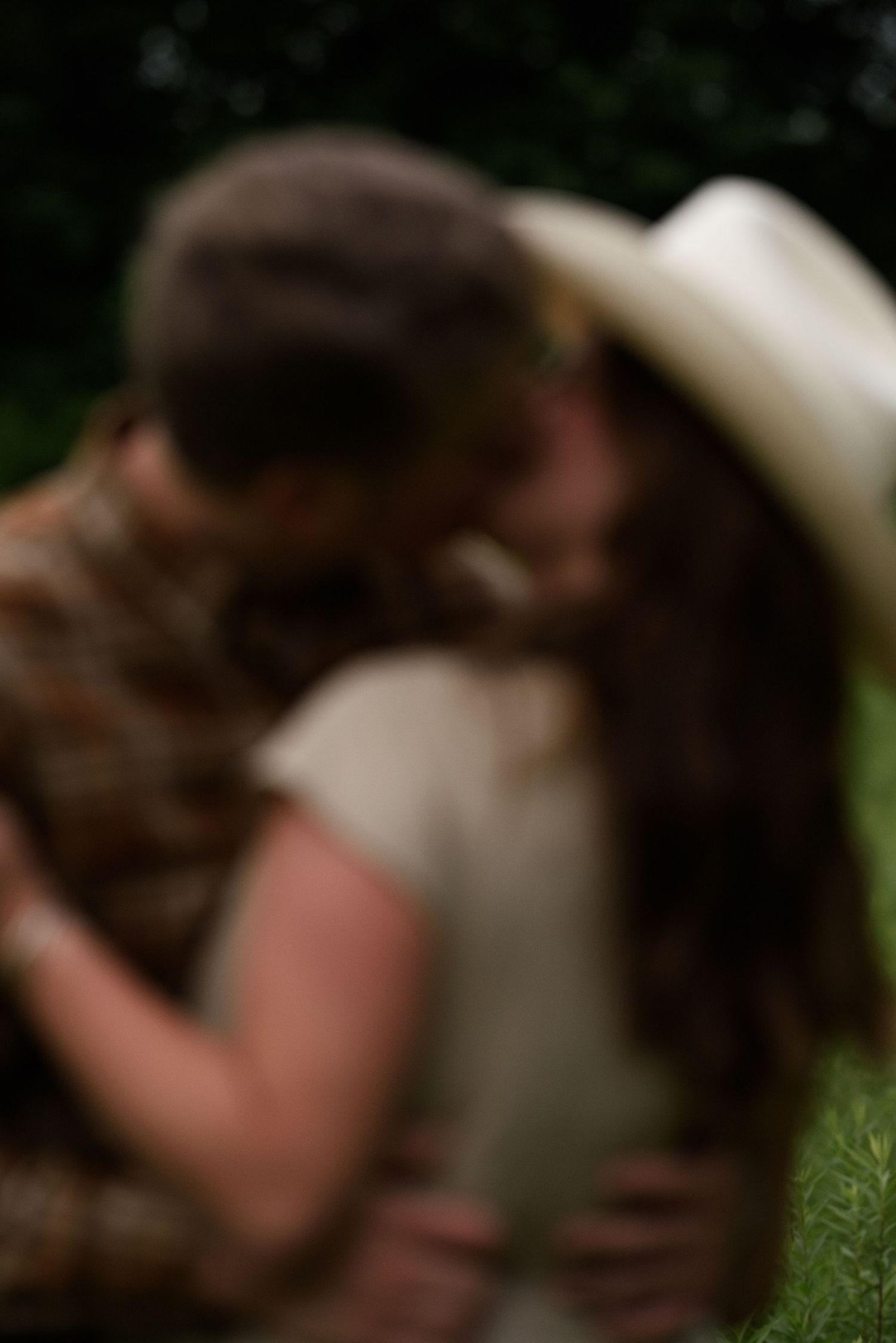 Blurry romantic couple kissing outdoors, woman wearing white hat, embracing in green nature setting.