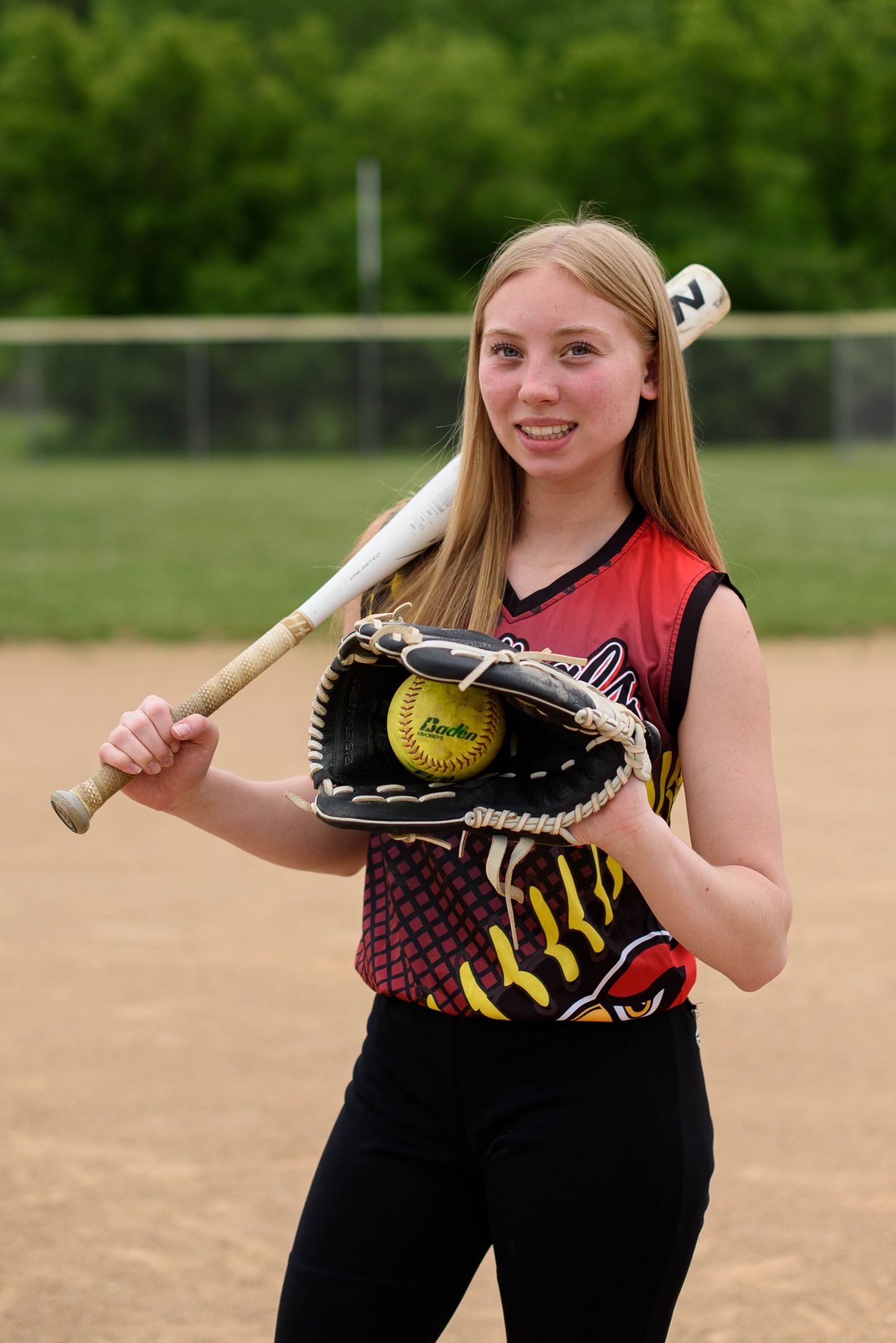 Young softball player poses on field holding bat and glove with yellow ball, wearing red and black team jersey.