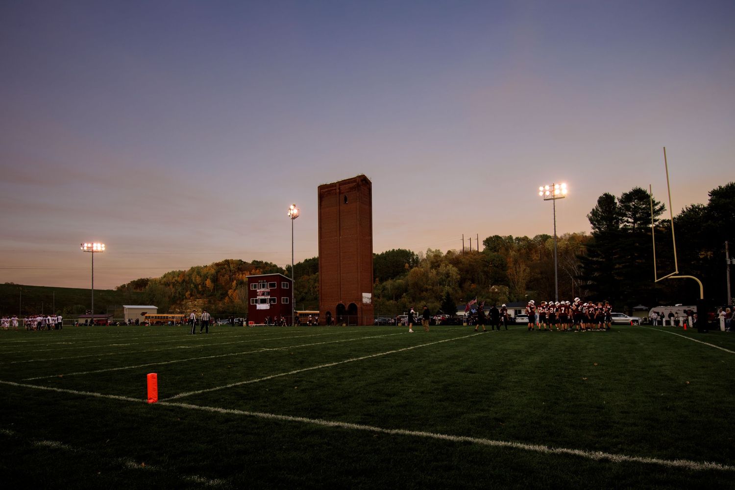 Football field at dusk with stadium lights, players gathered near end zone, brick tower landmark in background.