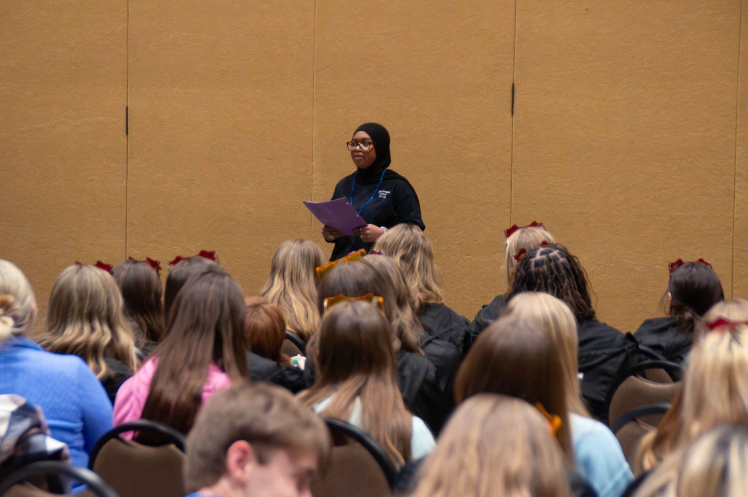 A speaker in a hijab addresses a large audience of students at a leadership conference.