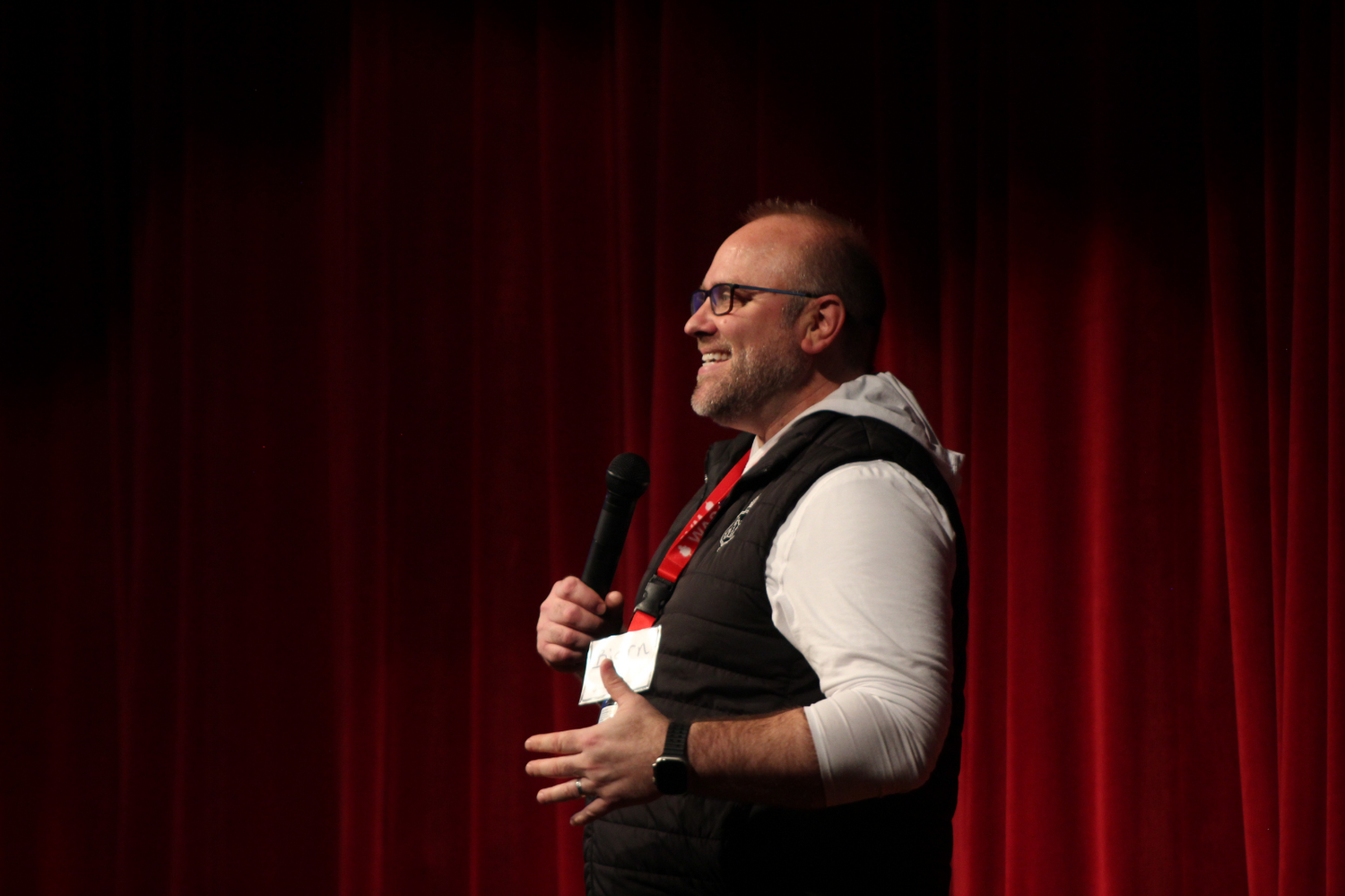 A bald man with glasses speaks into a microphone on stage in front of a red curtain.