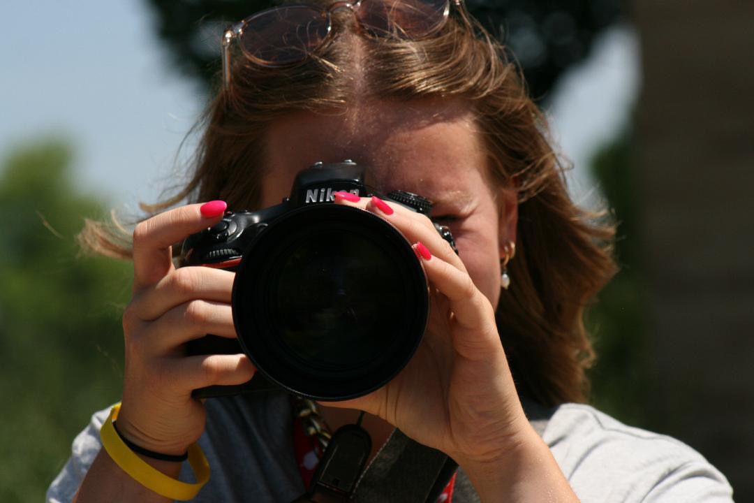 Woman with red nails holding a Nikon DSLR camera up to her eye, photographing outdoors on a sunny day.