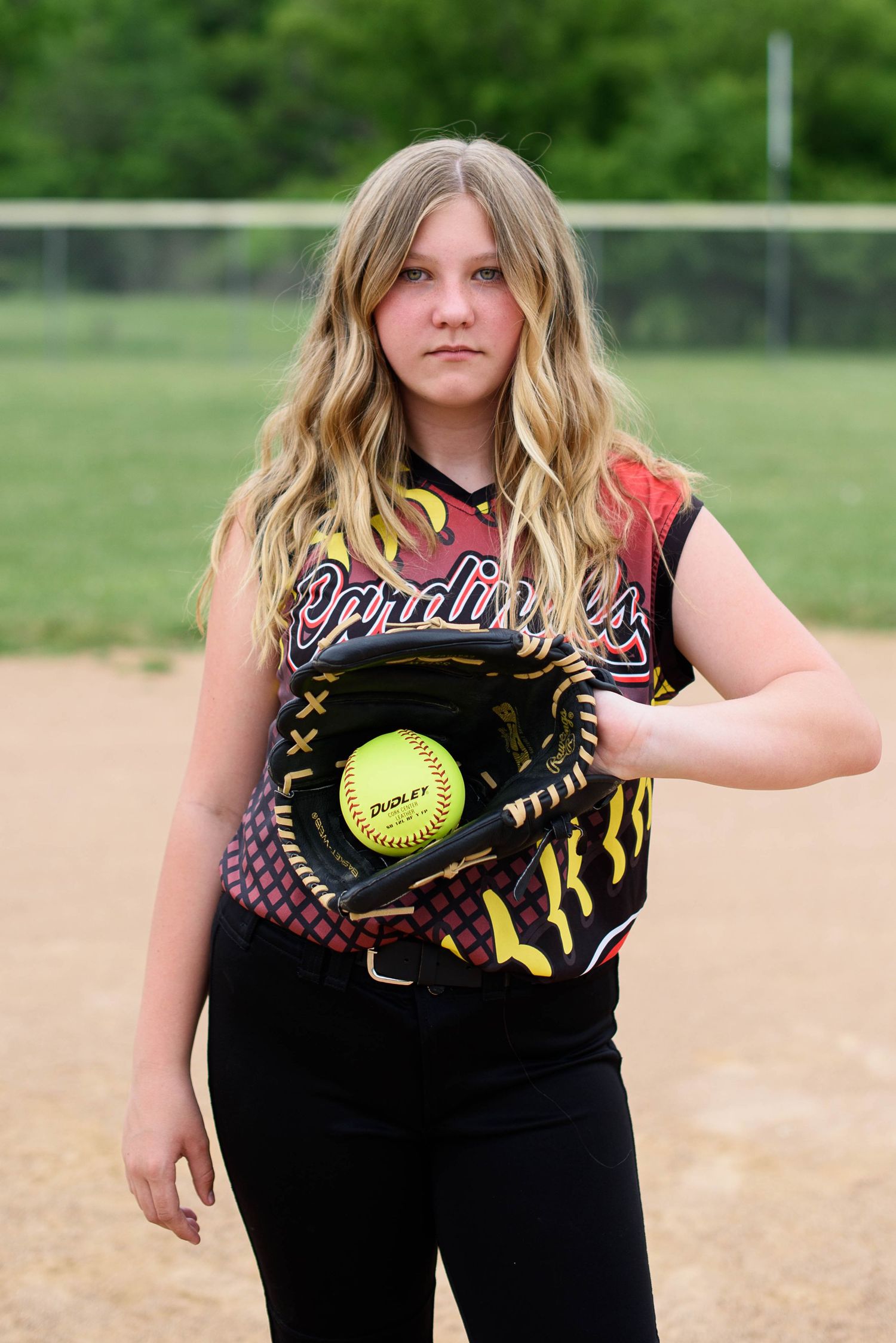 Young female softball player in Cardinals jersey holding glove with yellow ball on dirt field.
