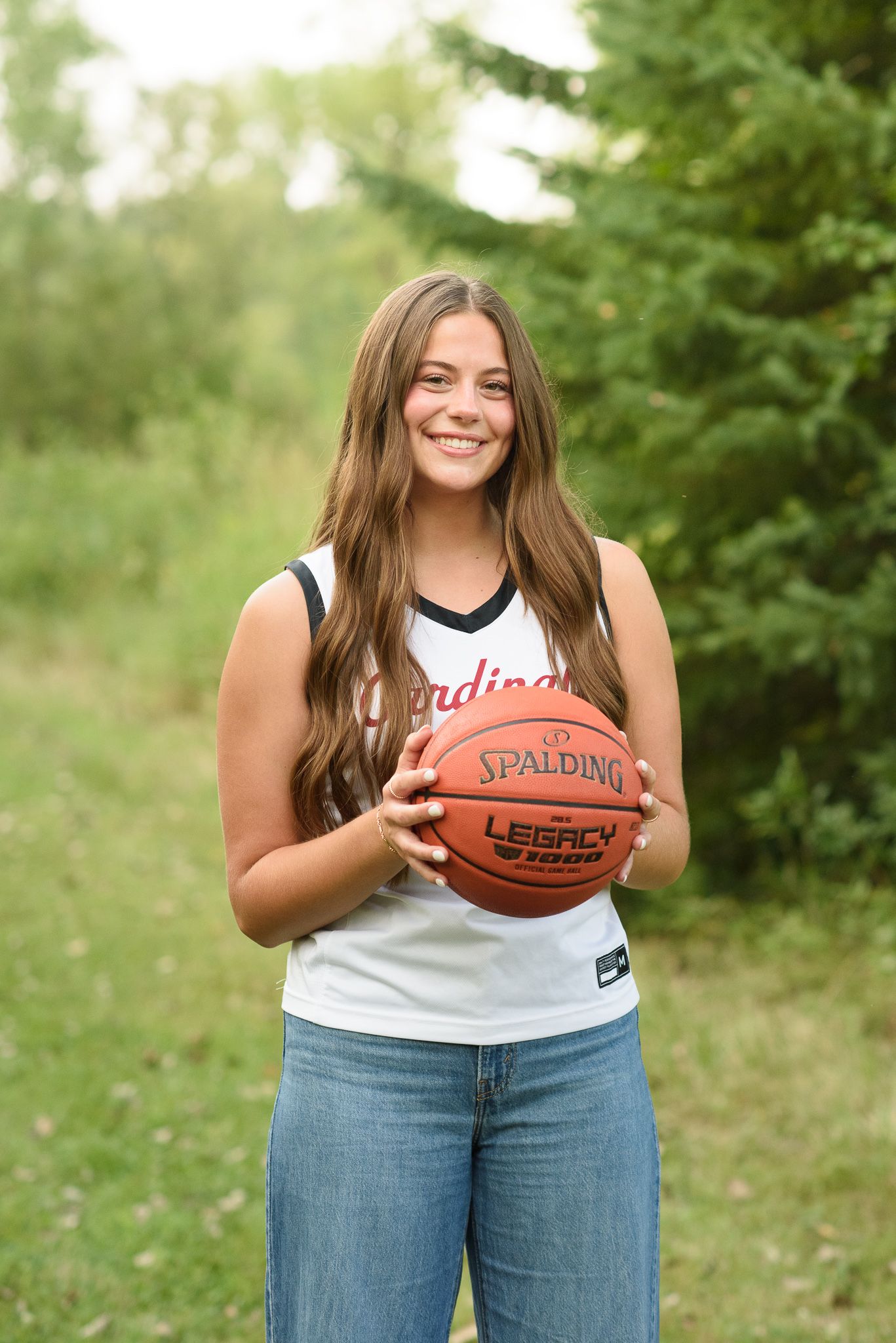 Smiling high school senior girl holding Spalding basketball wearing Cardinals white jersey outdoors.